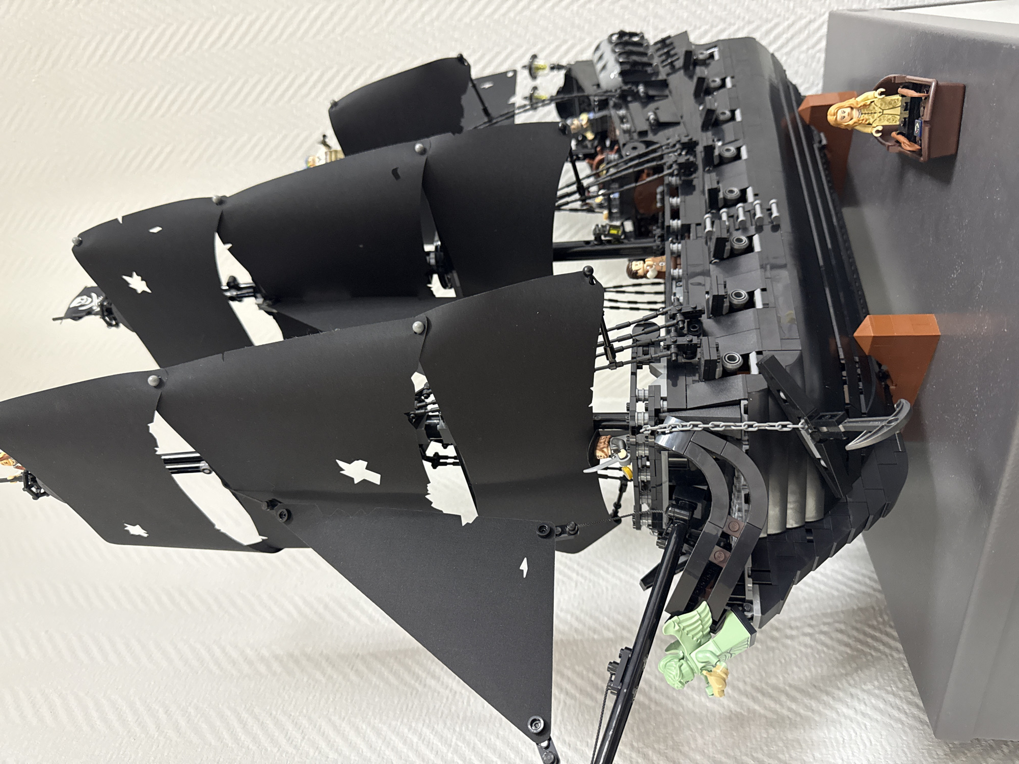 The image shows a detailed Lego model of a pirate ship with black sails mounted on a gray surface. The ship is positioned on the edge of the surface with part of the stern supported by brown Lego blocks. A Lego minifigure is placed in a brown Lego structure attached to the gray surface, near the ship's stern. The sails display stylized cuts and shapes, possibly representing damage or the ship's design. There is no indication of Krasnoyarsk, Russia, in the image.