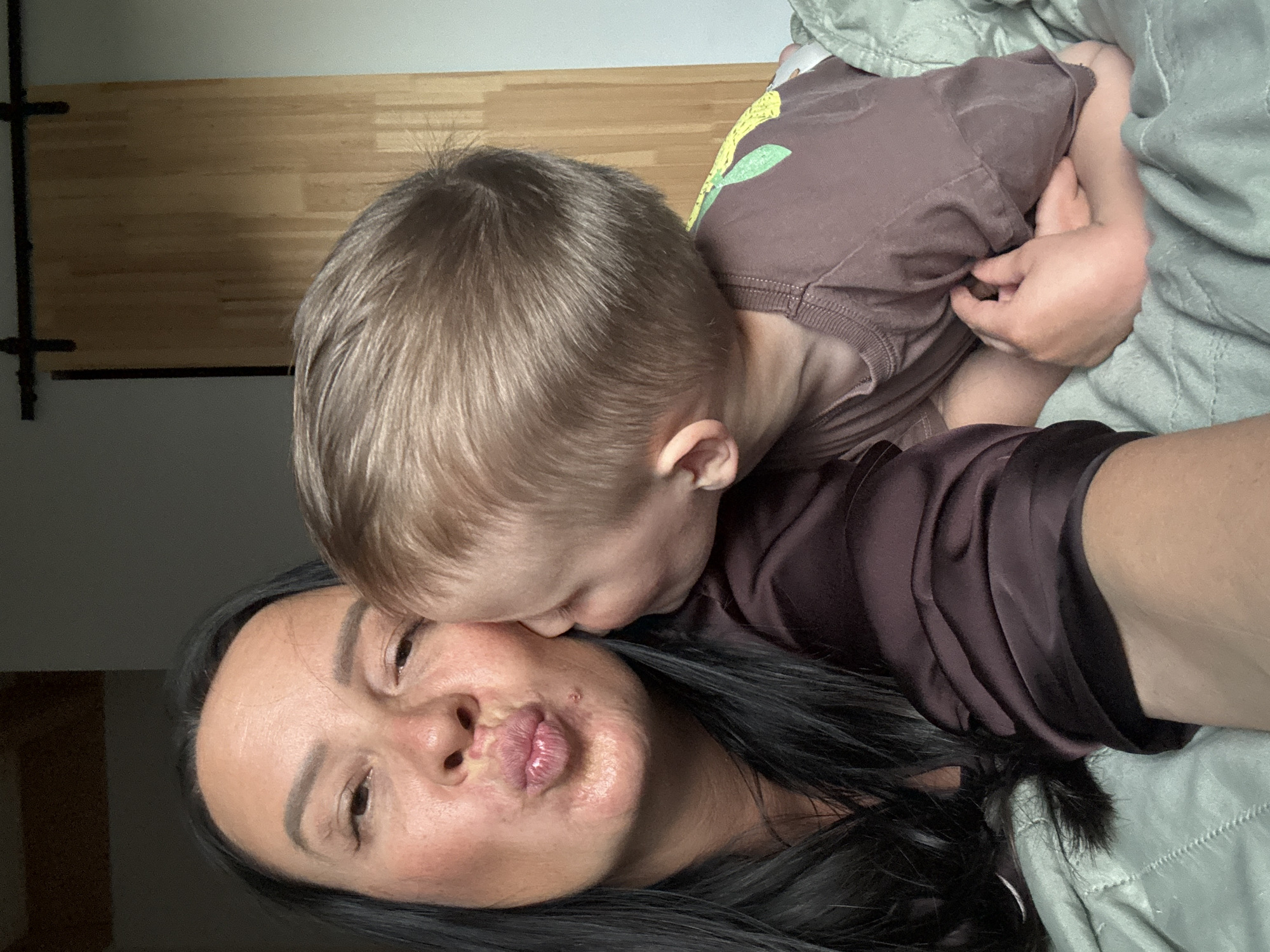 A selfie depicts an adult female with long black hair and a child with blond hair. The child is leaning in to kiss the adult female's cheek. The background includes what appears to be a wooden wall and part of a bed with a gray blanket. The location and specific city are not determinable from the visual information provided.