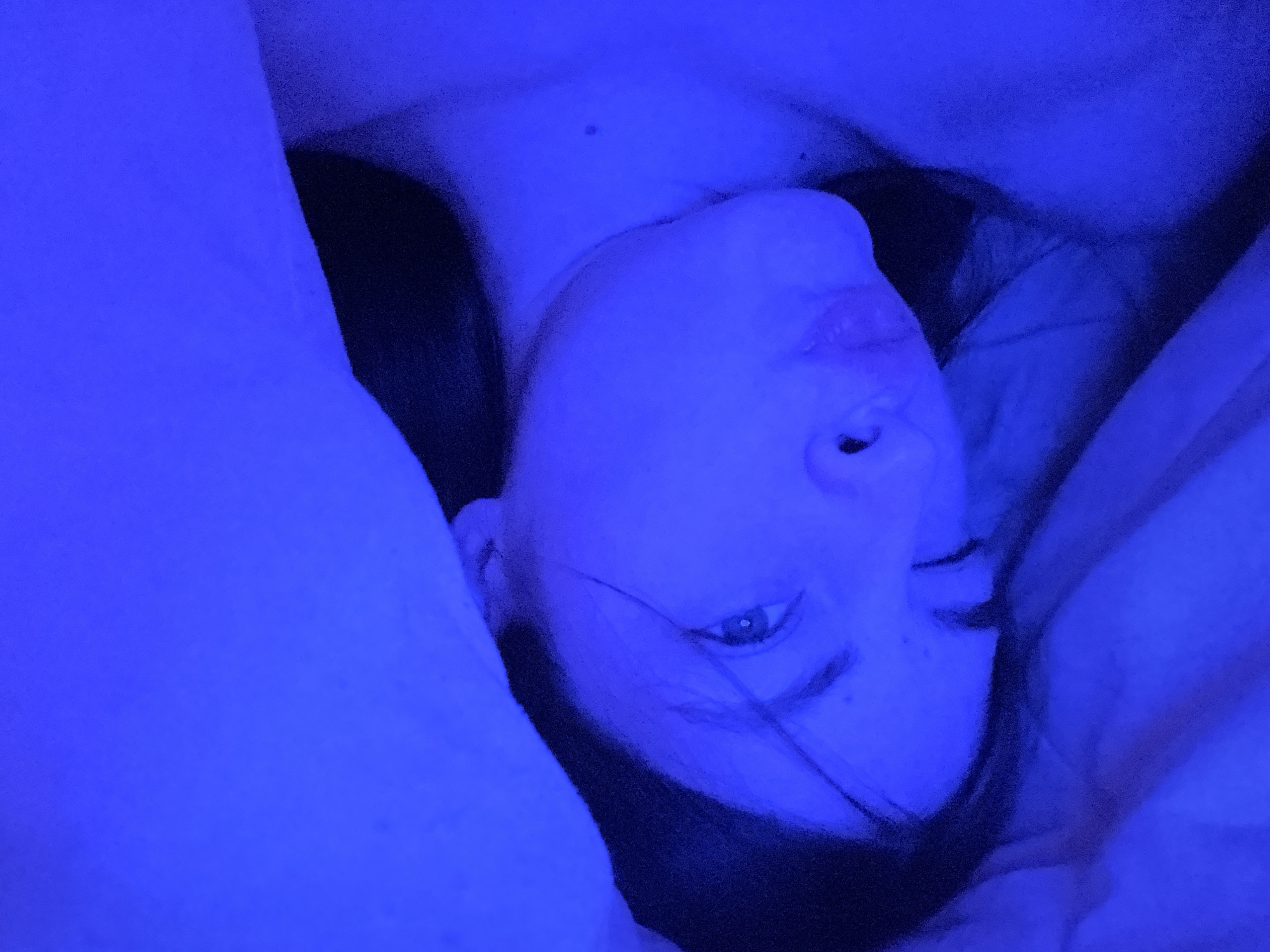 An overhead shot captures a person, likely a young adult or teenager, lying down in an indoor setting, possibly a bedroom. The entire scene is bathed in an intense, artificial blue light, giving the image a moody or stylized appearance. The subject's head is tilted, with their face looking upwards towards the camera. One eye is clearly visible and open, while the other appears partially closed or squinting. Their mouth is slightly open, revealing the lips. Dark hair frames their face, blending with the shadows created by the blue light.
The person appears to be surrounded by bedding or blankets, which are also illuminated by the strong blue light, showing soft folds and textures. The dominant blue hue obscures natural skin tones and casts deep shadows, particularly around the sides of the person's head and neck. The overall impression is one of relaxation or a candid moment captured under specific lighting conditions. There is no visible text in the image. The location is an indoor space, consistent with the provided context of Saint Petersburg, Russia, although no specific landmarks are visible.