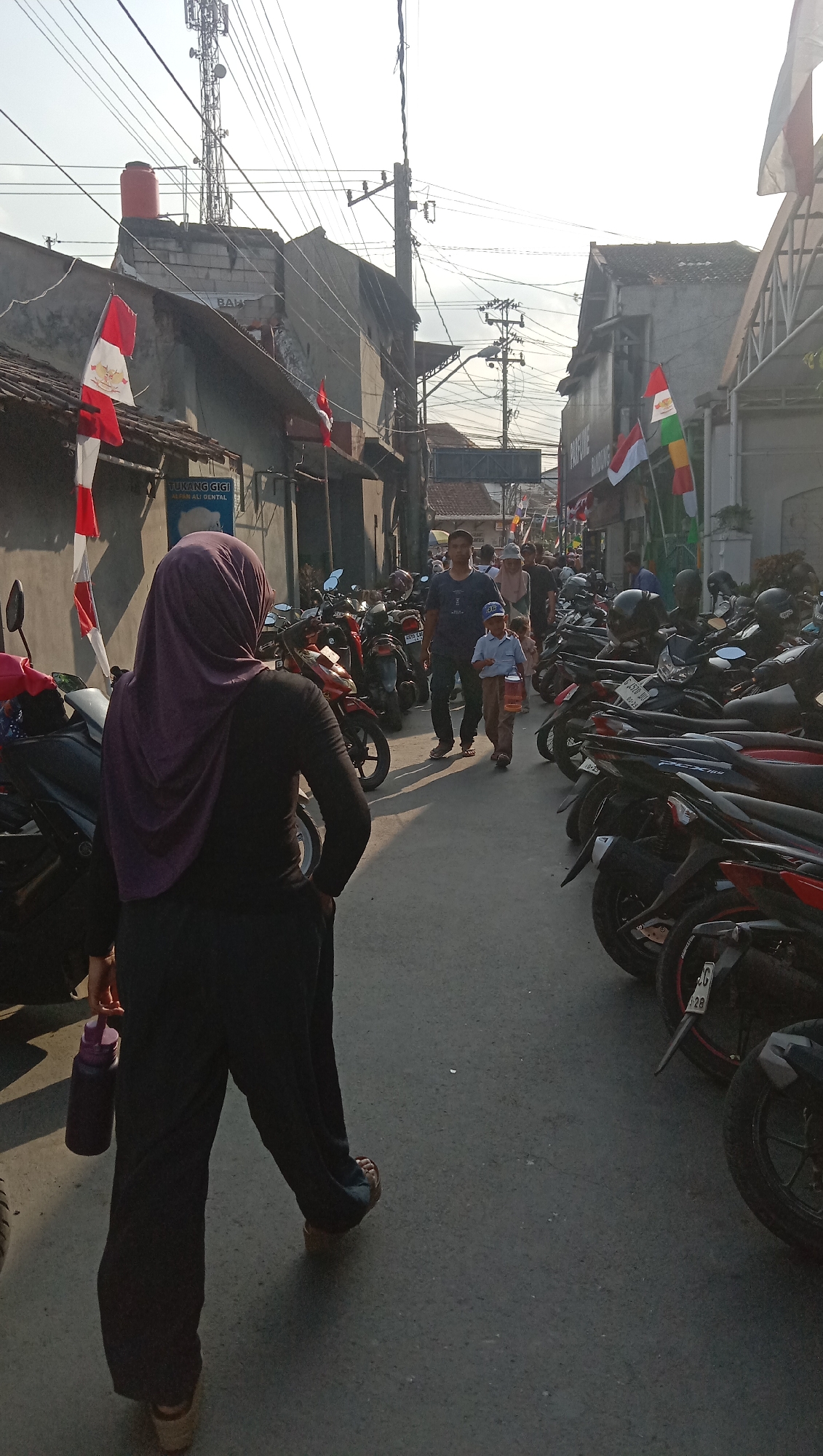 The image captures a street scene in Pesantren, Indonesia, on a bright, sunny day. The street is lined with buildings and densely packed with motorcycles, indicating a busy urban environment. A woman wearing a purple hijab and dark clothing is walking away from the camera, holding a dark water bottle in her left hand. Further down the street, a man in a dark t-shirt and dark pants, accompanied by a young boy in a light blue shirt, a cap, and brown pants, is walking towards the camera. The boy is carrying a small cage. Several Indonesian flags are visible, attached to buildings and poles, suggesting a national celebration or event. The overall atmosphere is one of everyday life in a bustling Indonesian city.