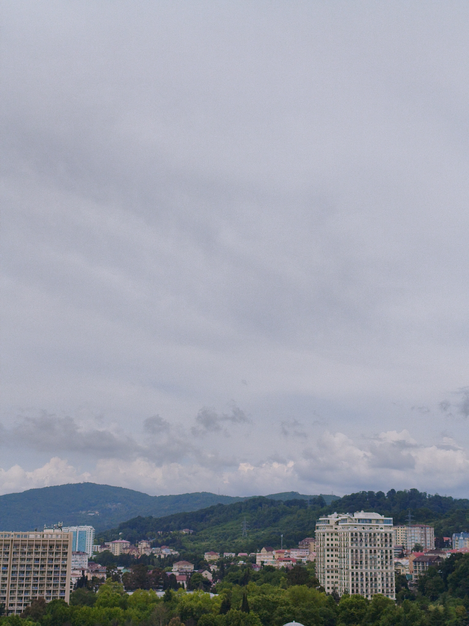 The image shows a cityscape in Sochi, Russia, with a focus on a cluster of buildings nestled against a backdrop of lush green hills. In the foreground, a tall, light-colored apartment building with numerous balconies dominates the left side of the frame. To its right, a slightly shorter, modern apartment building with a white facade and a distinctive rounded roof structure stands out. Behind these buildings, a densely wooded hillside rises, dotted with smaller houses and structures, suggesting a suburban or semi-rural area. The sky is overcast with a soft, diffused light, creating a somewhat muted color palette. The scene appears to be a residential area with a mix of modern high-rises and older, more traditional homes, set against a natural landscape.