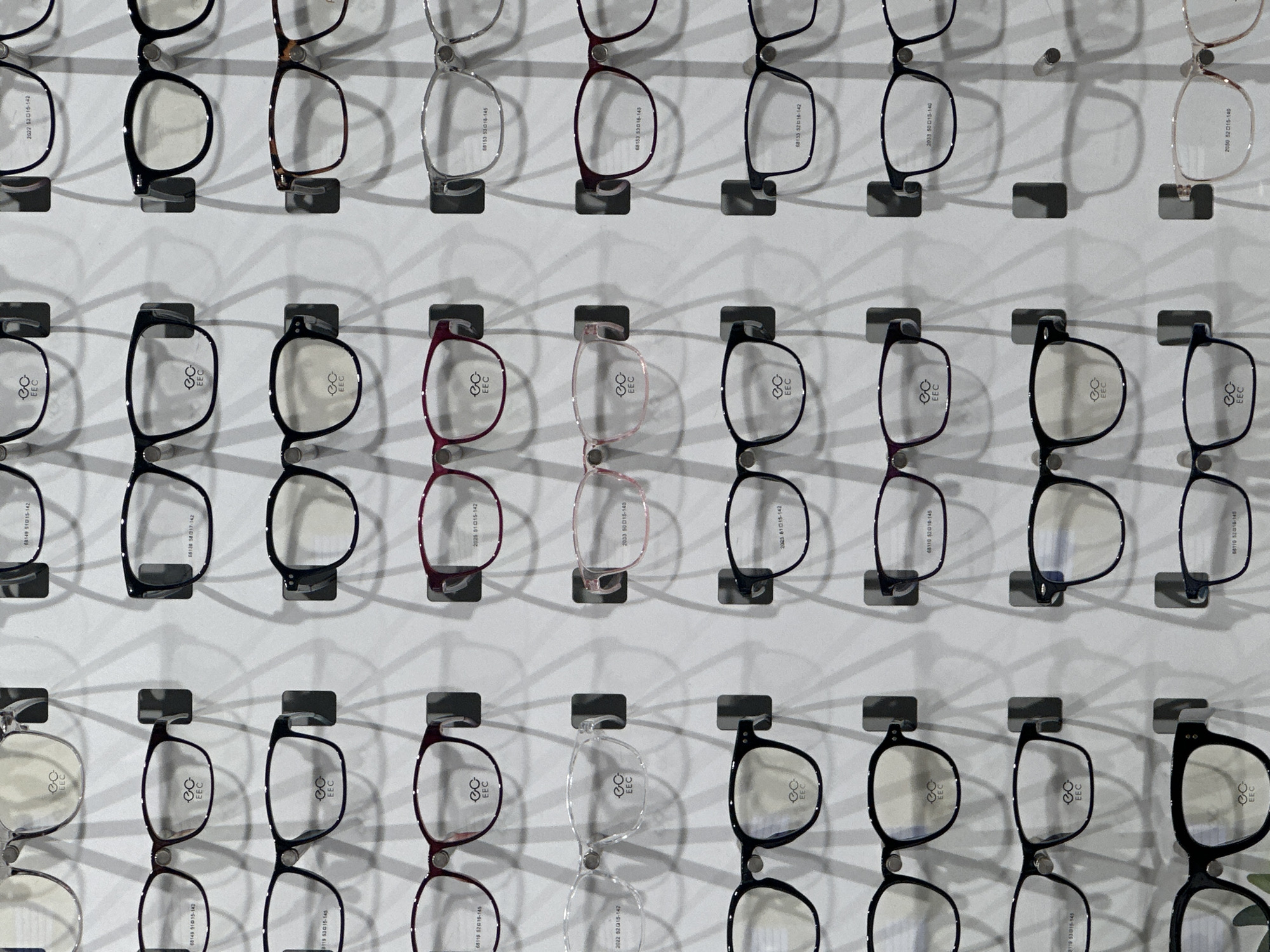 The image shows a display of eyeglasses on a white wall, likely in an optician's shop or a similar retail setting. The glasses are arranged in rows and columns and are of varying frame styles and colors, including black, silver, and shades of red. Each pair of glasses is mounted on a small rectangular mount. There are inscriptions, including the text "EEC" and model numbers like "68149 51015-142", printed on the glasses. The lighting appears to be bright and even.