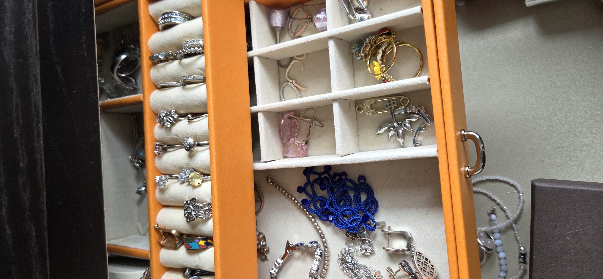 The image shows a close-up, eye-level view of an open, orange jewelry box. The box has multiple compartments, each lined with a cream-colored fabric. The top row of compartments contains an assortment of rings, necklaces, and earrings. The bottom row of compartments holds more rings, a blue crocheted item, a pink faceted stone with a safety pin, and some other assorted jewelry pieces. The jewelry box is placed on a light gray surface, and a portion of a dark brown box is visible in the lower left corner. The background is dark, suggesting the interior of a room, possibly a dresser or closet. There are no people or animals visible in the image. The lighting is even and appears to be artificial, with no discernible shadows. The overall scene is static and still.