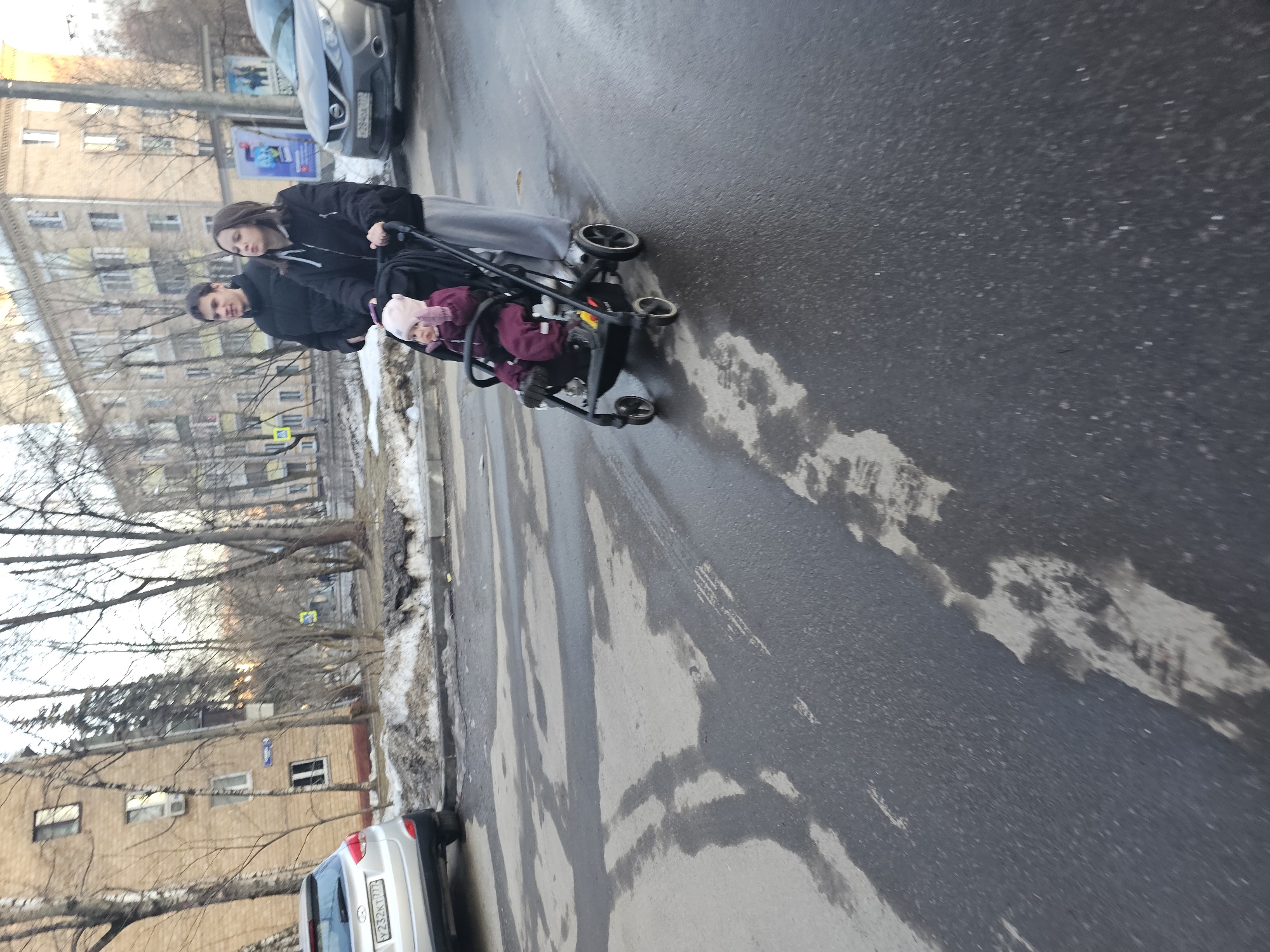 The image displays an outdoor street scene on Ulitsa Lavochkina in Khimki, Russia. A young adult female, dressed in a black hooded jacket, light grey sweatpants, and white sneakers, pushes a black child's stroller. Seated in the stroller is a toddler wearing a pink hat, maroon outerwear, and dark footwear. A young adult male, in a black puffer jacket, walks to the right and slightly behind the female. They are situated on a wet, dark asphalt road exhibiting patterns of moisture and faint tire marks. Along the road's edges, accumulations of dirty snow and slush are visible. In the background, numerous bare deciduous trees line the street, interspersed with multi-story residential buildings featuring brick and light-colored facades. A silver Hyundai Tucson SUV (Russian license plate Y232KT777) is parked on the left side of the road, partially on a snowy verge. Further down the street on the right, a grey Nissan Qashqai SUV (Russian license plate O280CA777) is parked. A blue street sign indicating "ул. Лавочкина" is partially visible on a pole near the left-parked vehicle. The scene is illuminated by ambient daylight under what appears to be an overcast sky.