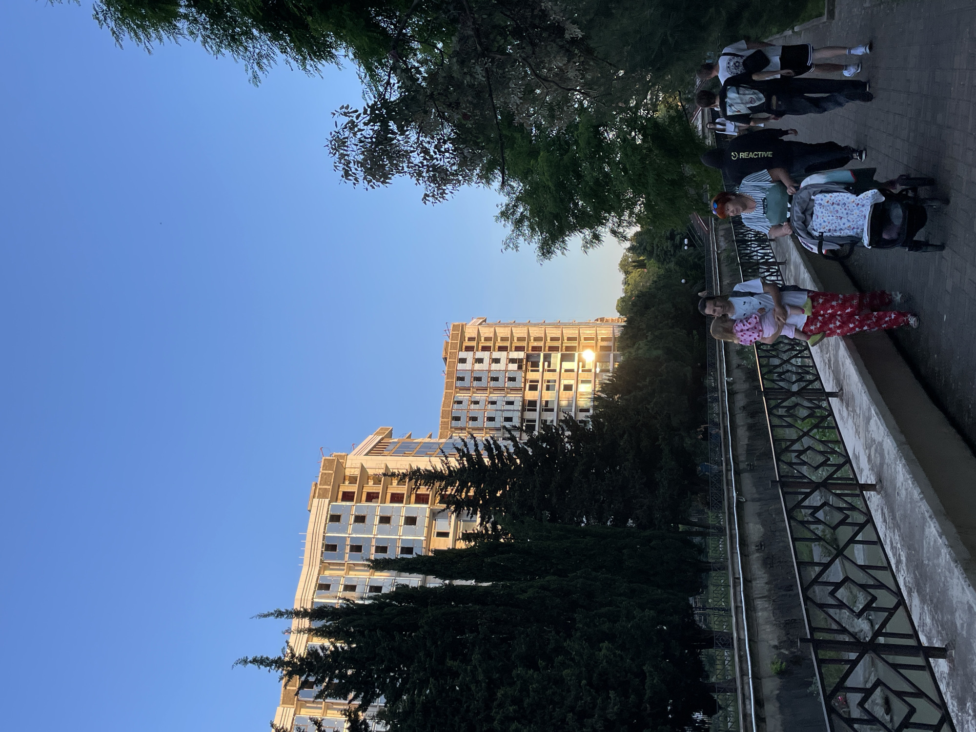 The image depicts an outdoor scene in Sochi, Russia, during late afternoon or early evening, evidenced by a clear blue sky and sunlight reflecting off distant structures. A paved pathway runs horizontally across the lower third of the frame, accommodating several individuals.
To the left of the pathway, a concrete-lined canal or river flows, bordered by a dark metal railing with a repeating diamond pattern. Beyond the canal and behind a row of tall, dark green coniferous trees, two multi-story buildings are under construction. These buildings feature exposed concrete frameworks and numerous installed reflective blue-tinted windows. The setting sun creates a bright reflection on the lower windows of the building on the right.
On the pathway, a group of people walks away from the viewer. From left to right:
1. A man in a white t-shirt and red-and-white patterned pants holds a young child (toddler) dressed in a pink top and light-colored bottoms. The child's head is turned to the left.
2. A woman with reddish hair, wearing a striped t-shirt and dark overalls, pushes a dark-colored baby stroller that contains a patterned blanket.
3. A person wearing a black hoodie with "REACTIVE" written in white on the back, and dark pants.
4. Two young men. The one on the left wears a dark t-shirt with a graphic design, dark pants, and carries a dark shoulder bag. The one on the right wears a white t-shirt with a graphic design, dark shorts, and also carries a dark shoulder bag.
Further along the path, additional human figures are faintly visible in the distance. The environment combines natural elements like trees with urban infrastructure and ongoing development.