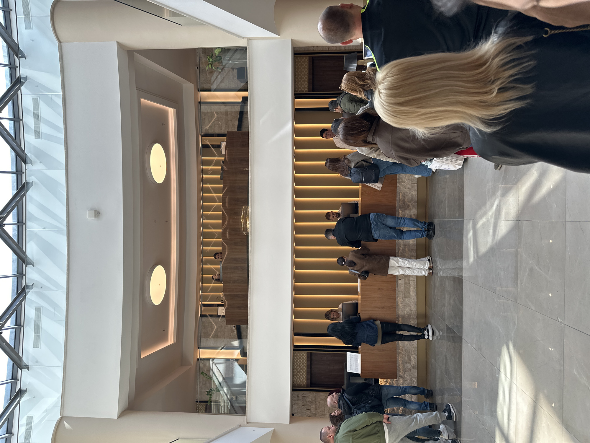 This image depicts a modern and spacious indoor area, likely a hotel lobby or a large reception hall, set in Kazanlak, Bulgaria. The primary focus is a queue of numerous people extending towards a long, contemporary reception desk on the right side of the frame. Most individuals in the line are seen from behind or the side, appearing to wait to check-in or for assistance. Staff members are visible behind the wooden reception desk, attending to guests, with computer monitors also on the desk.
The architecture is characterized by high, white walls and ceilings, clean lines, and extensive use of glass. A grand staircase with wooden steps and integrated strip lighting ascends in the background, reflected prominently in a large glass partition. Natural light floods the space from tall windows or skylights on the left, illuminating the light-colored, possibly tiled or marble, floor and casting soft shadows. A large, circular light fixture is mounted on an upper wall. Potted plants add a touch of greenery, visible through glass elements. The overall ambiance is bright, orderly, and modern. No discernible text is visible within the frame.