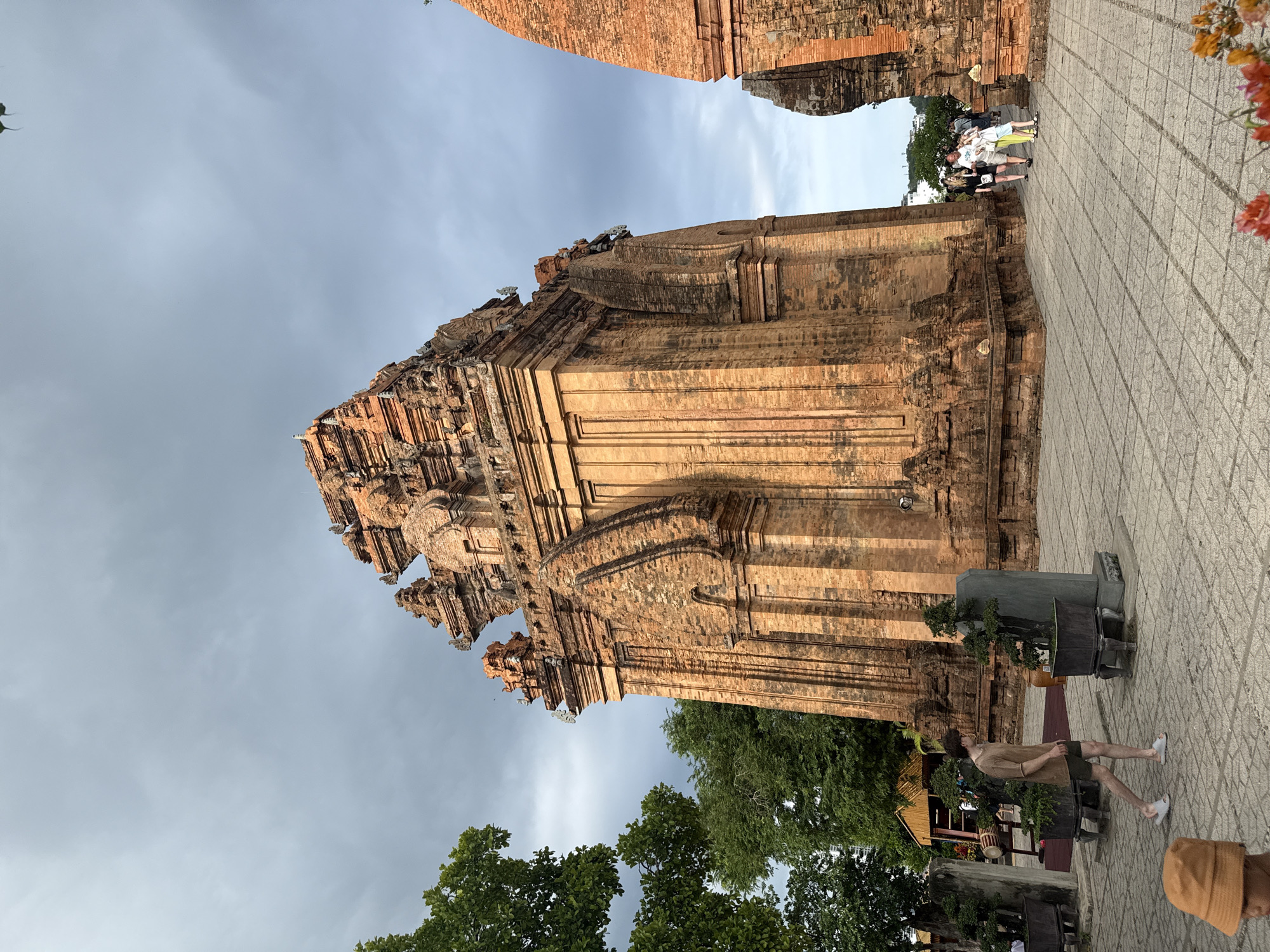The image depicts the Po Nagar Cham Towers, a historical complex located in Nha Trang, Vietnam. The foreground shows a paved courtyard with several people. A young man in a brown t-shirt and shorts walks across the courtyard from left to right. To his left, a person wearing an orange bucket hat is partially visible. In the background, the distinctive red brick Cham towers dominate the scene, with intricate architectural details and weathered surfaces. Several other people are seen walking near the towers in the middle ground. Lush green trees surround parts of the complex, and the sky above is overcast with dramatic clouds. The overall impression is of a historical site with visitors exploring the ancient structures.