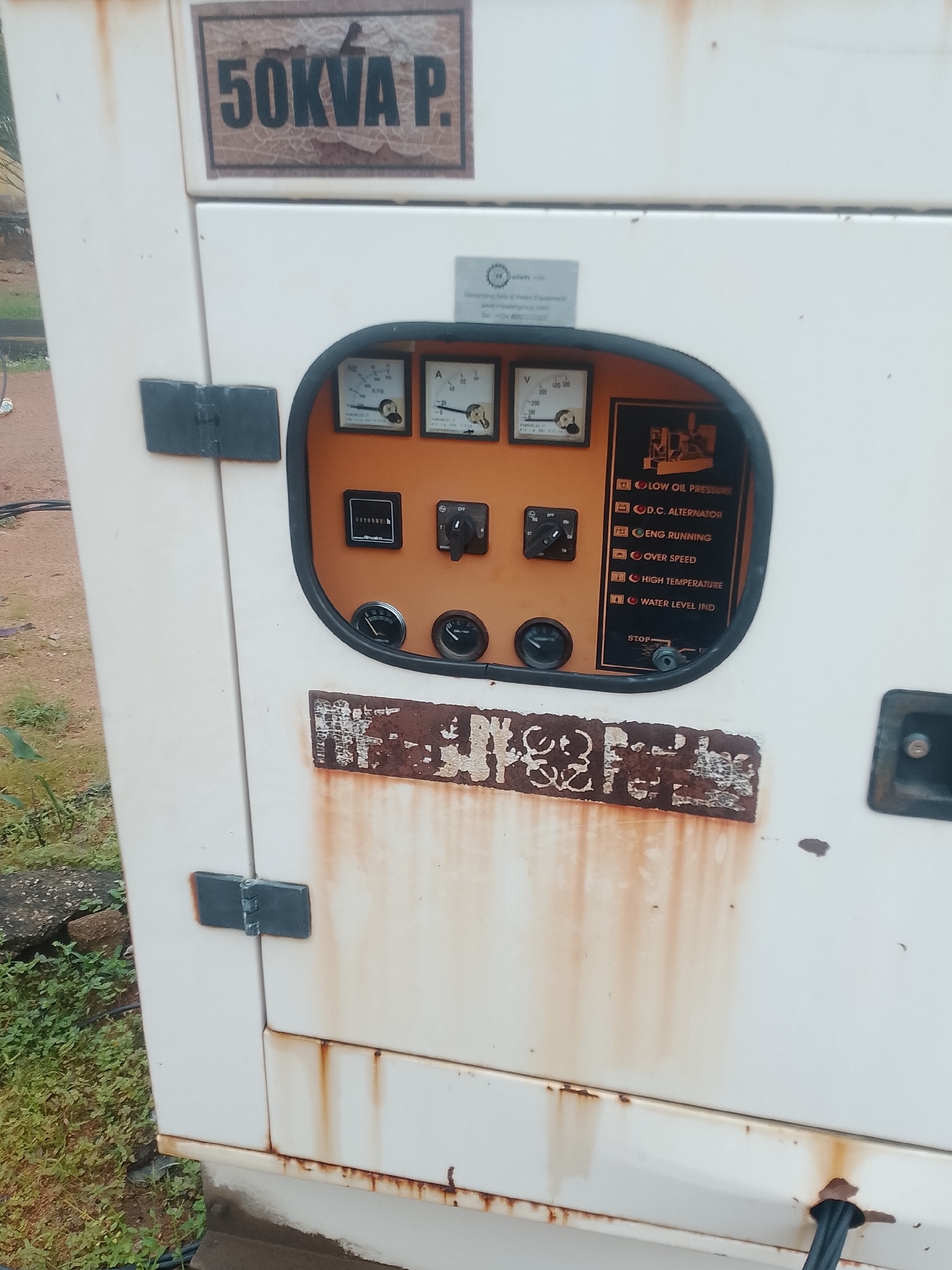 The image shows a close-up of a weathered, white generator unit. The top of the unit features a label that reads "50KVA P." with a cracked, brown backing. Below this, a rectangular panel contains several gauges and indicators. There are three circular gauges visible, two with needles pointing to readings, and one with a needle at approximately the 10 o'clock position. A digital hour meter displays "137059.2 h." To the right of these is a control switch with positions labeled "OFF," "T," "R," and "RN." A black rectangular panel to the right of the gauges contains several indicator lights and labels, including "LOW OIL PRESSURE," "D.C. ALTERNATOR," "ENG. RUNNING," "OVER SPEED," "HIGH TEMPERATURE," and "WATER LEVEL IND." A red "STOP" button is also present. A large, faded, and partially obscured label with white lettering on a dark background is affixed to the lower front of the generator. The generator exhibits significant rust stains, particularly on the door and around the control panel. The unit is outdoors, with green foliage and a dirt path visible in the background. A black electrical cable emerges from the bottom right of the generator.