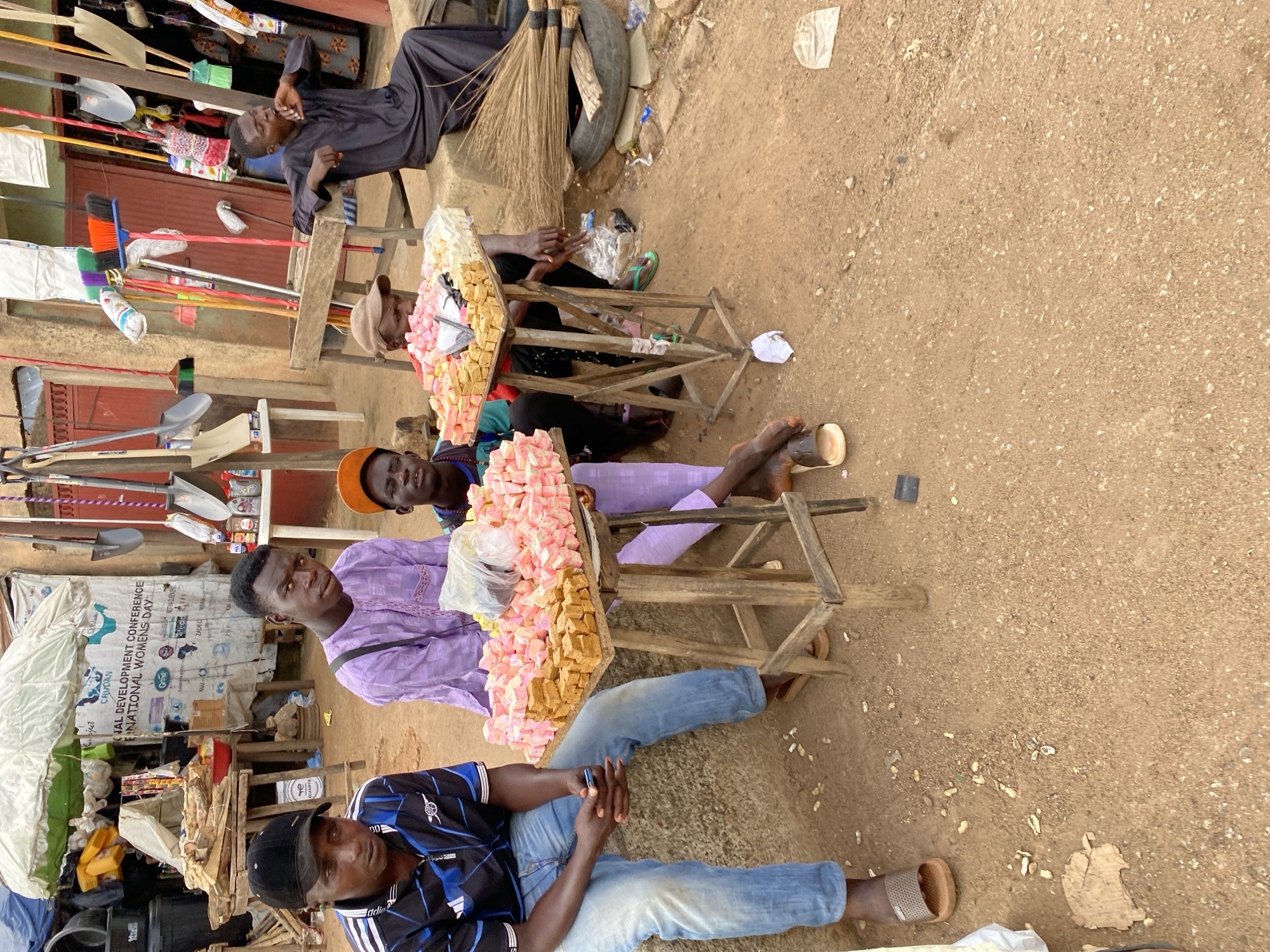 In Bukuru, Nigeria, a group of young men are gathered around stalls displaying colorful sweets. The scene takes place on a dirt ground, with various items scattered around, including brooms, a tire, and some discarded wrappers. In the background, a stall with a white tarp displays a banner that reads "NATIONAL DEVELOPMENT CONFERENCE INTERNATIONAL WOMENS DAY."
One young man, wearing a black baseball cap and a blue and black striped shirt, sits on a concrete ledge, looking down at his phone. Another young man, dressed in a purple traditional outfit, sits beside him, his gaze directed forward. In the center, a younger boy in an orange cap is seated and appears to be sleeping. A fourth young man, wearing a black traditional outfit, is perched on the edge of a concrete structure, yawning.
The stalls feature wooden legs and are topped with trays of sweets. The sweets are predominantly pink and yellow, neatly arranged. A plastic bag of sweets is placed on one of the trays. The overall impression is of a street-side market or gathering, with a focus on the display and sale of confectionery.