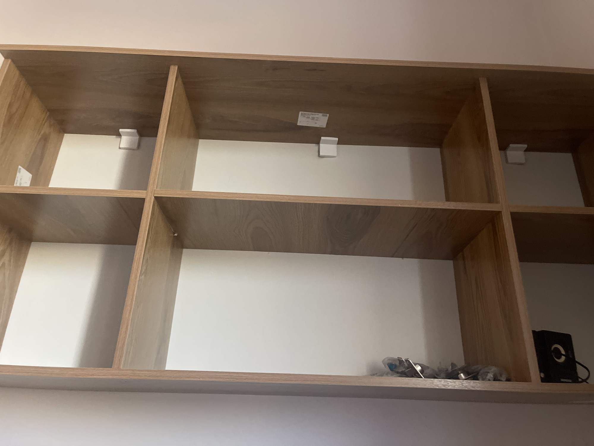 An empty, light-colored wooden shelf unit with six compartments is visible against a light-colored wall. Some compartments have white inserts. There are small white clips or brackets attached to the back of some compartments, and a small white sticker on the back of another. In the bottom right compartment, a small black electronic device with a circular opening and a wire is placed on the shelf, next to some other small objects wrapped in plastic. The scene appears to be indoors, possibly in a domestic or retail setting. The image does not contain any people or indicate a specific location in Qarshi, Uzbekistan.
