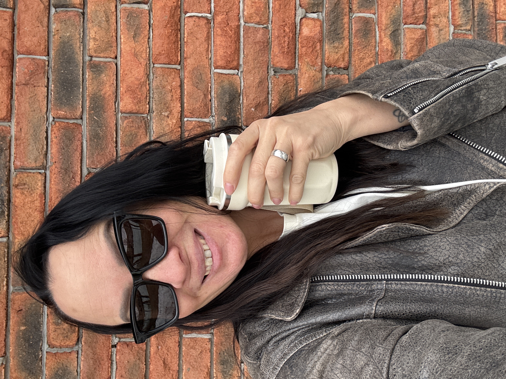 A woman is lying on a brick surface, possibly a wall, holding a white cup in her right hand. She is wearing sunglasses and a gray leather jacket over a white shirt. Her hair is long and dark. The background is a brick wall. The location cannot be determined from the image.