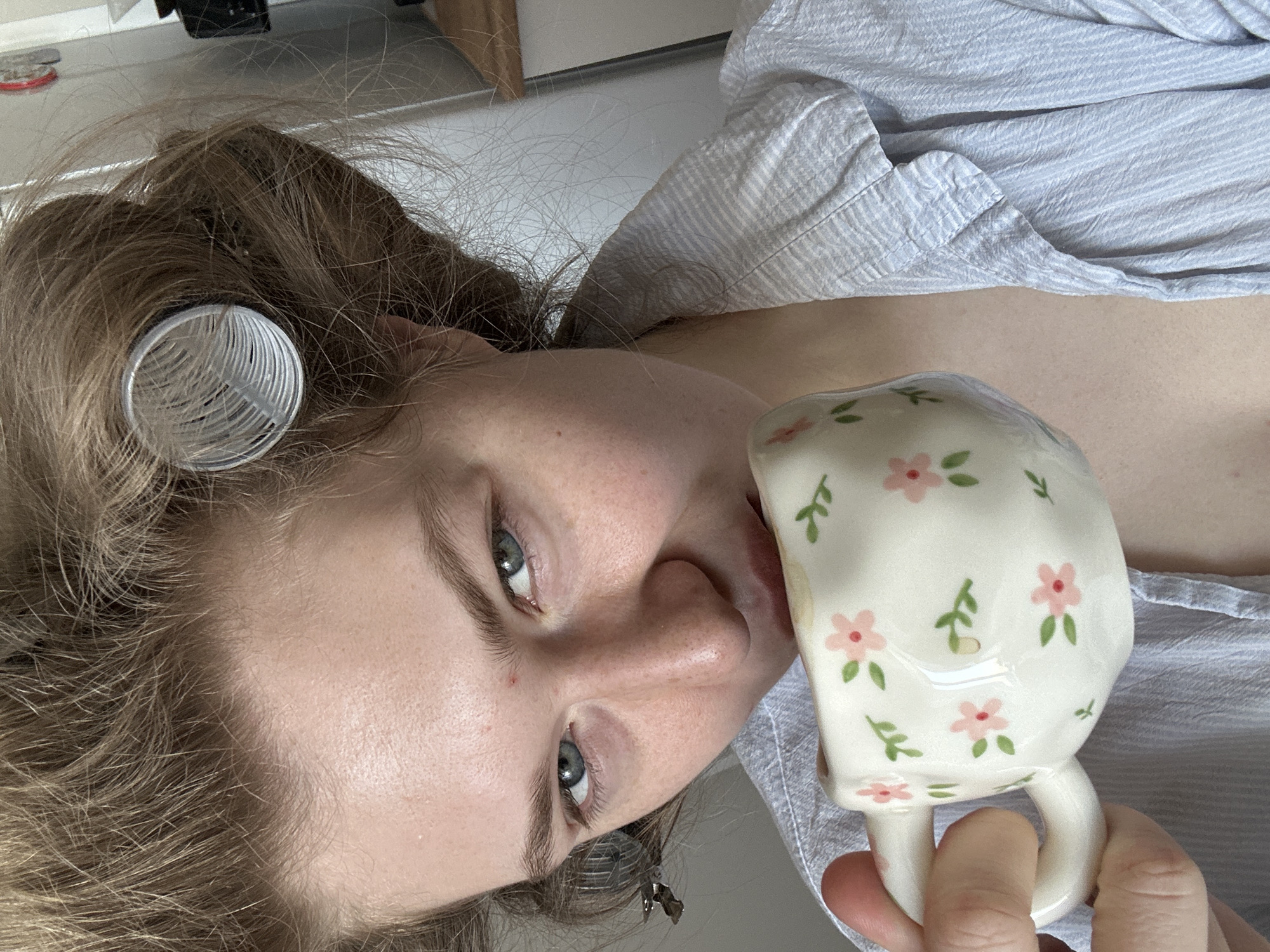 A young woman is shown in a close-up, slightly angled view, holding a floral-patterned mug close to her face, possibly drinking from it. She has light blonde hair with one visible hair roller on the left side. Her skin is fair, and she has light blue eyes. She is wearing a light blue striped shirt or pajamas. The background is a neutral color, possibly a wall. The location is not specified, and there is no information to suggest this was taken in Kazan, Russia.