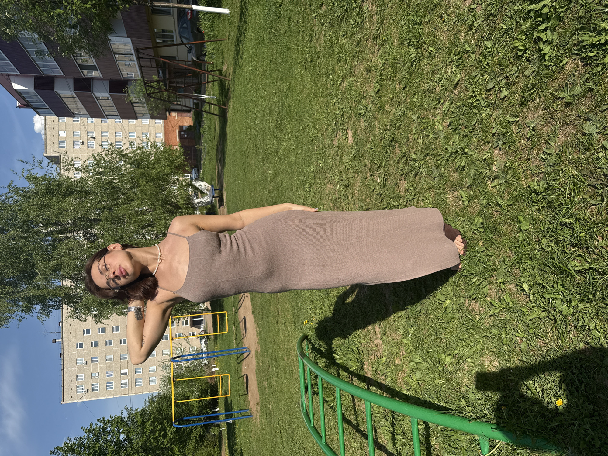 A young woman stands on grass in Sarapul, Russia. The environment includes green grass, a few scattered yellow dandelions, trees, playground equipment, apartment buildings, and blue sky with white clouds. The woman wears a brown dress, glasses, a pearl necklace, and a bracelet. She has dark hair. She touches her face with her left hand.