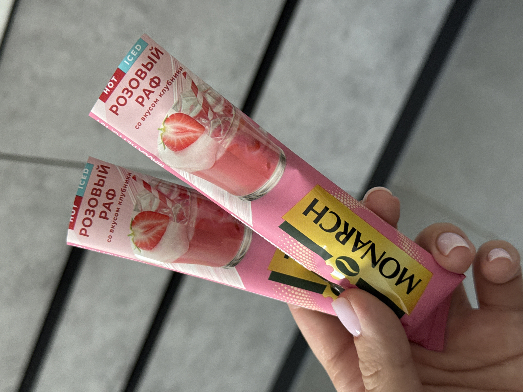 Two pink packages of "Monarch" instant coffee, labeled "ROZOVY RAF" (Pink Raf) with strawberry flavor, are held by a person's hand against a grey, textured background. The packages depict images of a strawberry, ice, and a pink beverage. The text "HOT" and "ICED" are also visible on the packages. The background appears to be a wall with horizontal paneling.