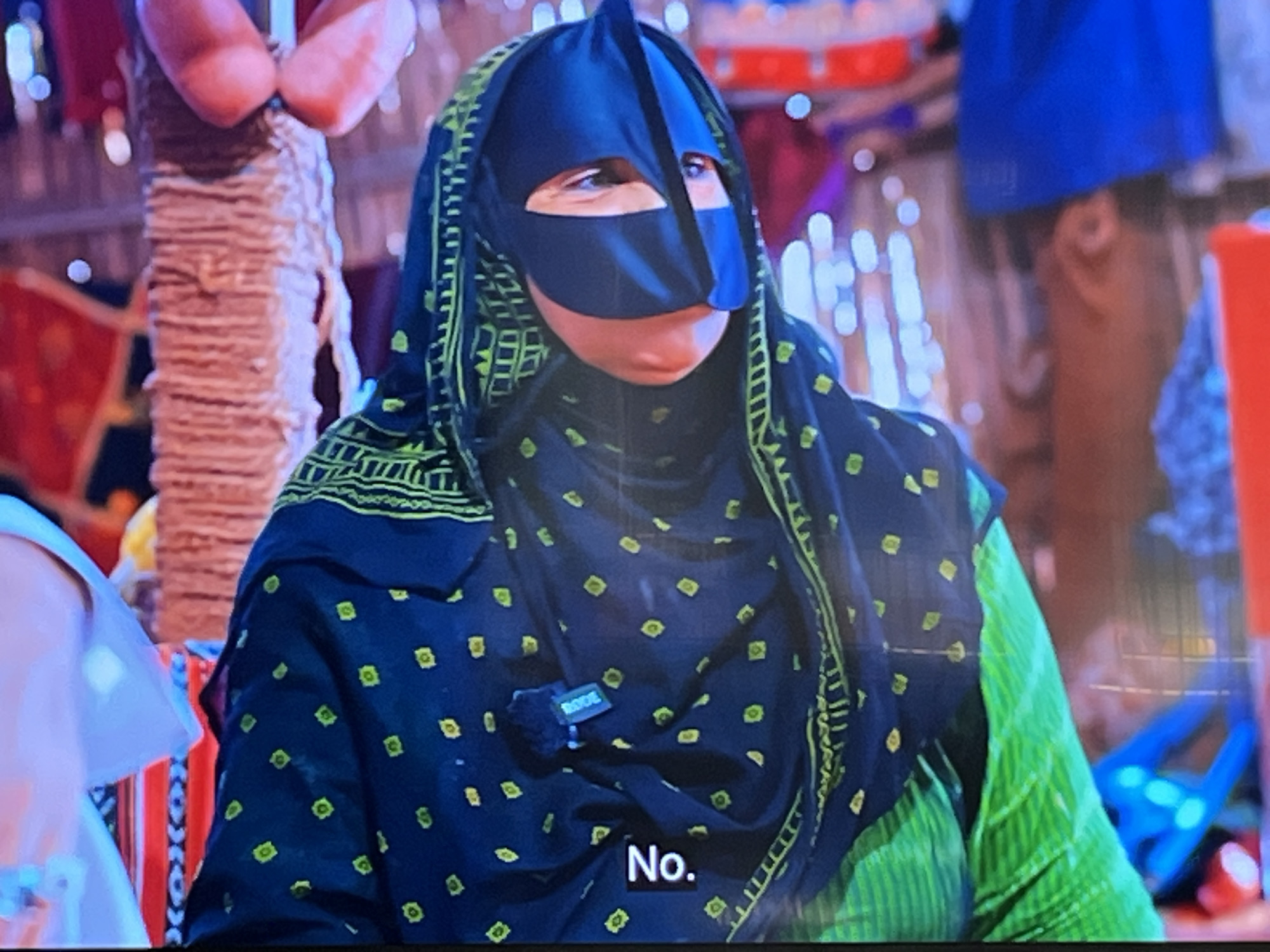 A person wearing a traditional niqab and headscarf is shown, with the niqab covering their face except for their eyes. The headscarf is black with a green embroidered border and small yellow diamond-shaped patterns. A microphone clip is attached to the fabric on their chest. A portion of their clothing underneath the headscarf appears to be bright green with vertical stripes. In the lower right corner of the image, the word "No." is displayed in white text on a black background. The background is blurred and appears to be indoors, with hints of decorative elements and other people or objects, but lacks distinct details. The context of Kharkiv, Ukraine, is not discernible from the visual information provided in the image.
