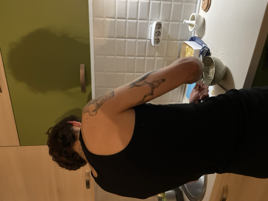 The image depicts an indoor kitchen scene in Boreti, Montenegro. A person, viewed from behind, is actively grating a light-colored substance into a grey ceramic bowl situated on a light-colored countertop. The individual wears a black tank top, revealing dark, curly hair and multiple tattoos: a bone-like structure on the upper left arm/shoulder, and an antlered skull design on the left bicep. To the person's right, a partially visible white box containing spaghetti or pasta is on the counter, alongside a white ceramic mortar and pestle. The kitchen features a white tiled backsplash with a textured, raised square pattern. Above the backsplash, the wall is comprised of light wood-grain upper cabinets with silver handles on the left and olive-green upper cabinets on the right. A prominent shadow, resembling a human head and shoulders, is cast on the olive-green cabinet. A white triple power outlet is mounted on the tiled backsplash on the right, and two small, dark wooden floating shelves are visible on the green wall section. A stainless steel sink is partially visible on the far left. The environment is illuminated by an unseen artificial light source.