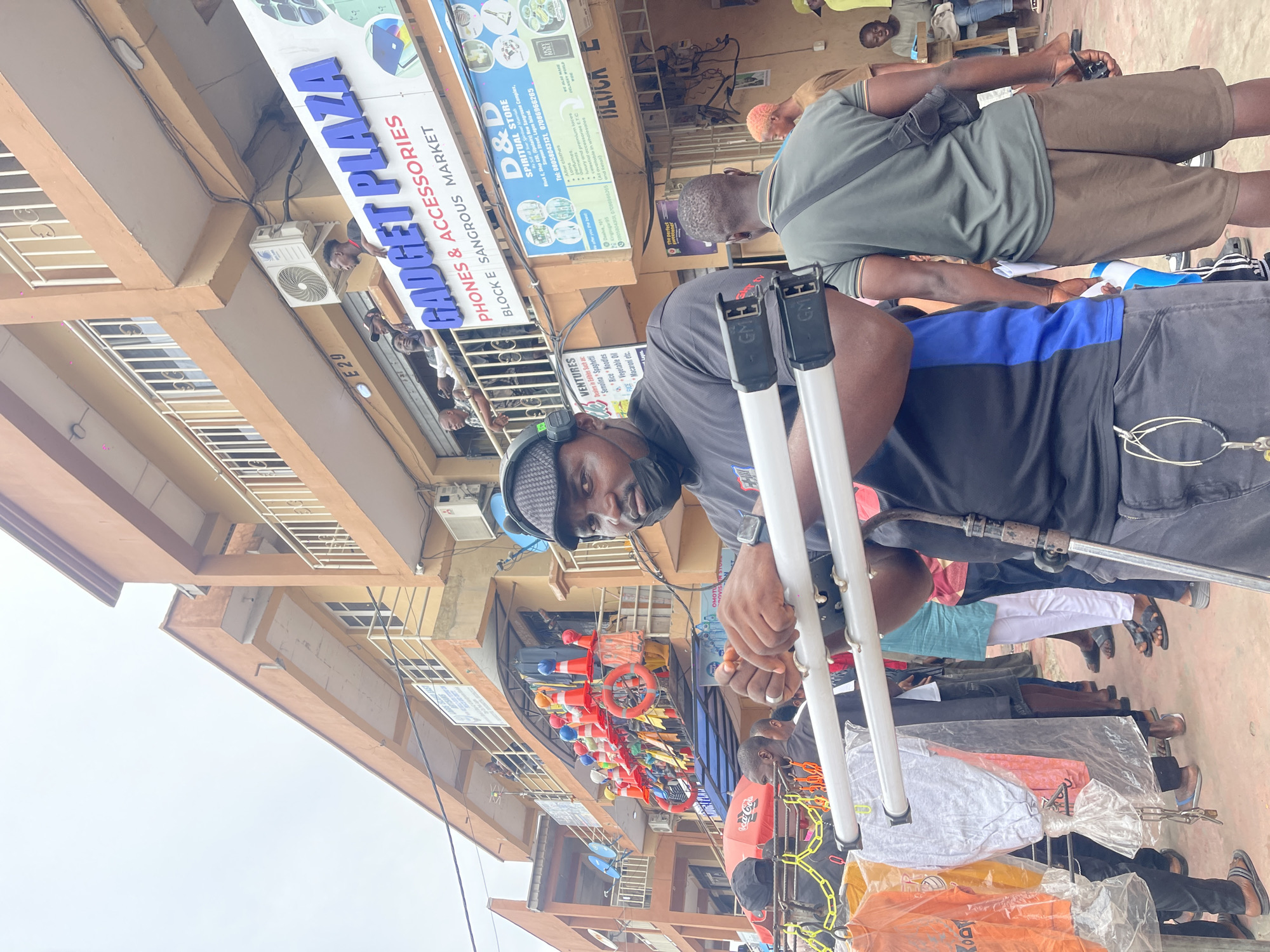 An outdoor, eye-level shot captures a vibrant street market scene, featuring a man in the foreground looking directly at the camera. He wears a black cap, a black face mask, a grey and blue short-sleeved top, and has a smartwatch on his left wrist. He is holding a white and grey metal frame, possibly a display stand, which has "GME" visible on its components. Various garments, including T-shirts, hang from a rack in front of him.
Behind the man, several other individuals are present, some walking, others standing. One man in a grey t-shirt and brown shorts is visible from behind. The background features multi-story buildings with balconies and railings, indicative of commercial establishments. Numerous signs are affixed to the buildings. A prominent sign reads "GADGET PLAZA PHONES & ACCESSORIES BLOCK E SANGROUS MARKET." Another sign identifies "D&D SPIRITUAL STORE Block E, Shop E30, (Upstairs) New Sangrouse Complex, Simpson Street, Lagos Island," listing items like "Alter clothes," "luxury and premium laces," and "celestial accessories." Further text identifies "VENTURES Dealers in Edibles Such as: Semolina Spaghetti, Rice Noodles, Vegetable Oil, Macaroni etc." Air conditioning units are mounted on the building facades. Decorations or merchandise, including red traffic cones and lifebuoys, hang from the upper level.
The lighting suggests an overcast daytime, contributing to an active, bustling atmosphere typical of a market street. The specific addresses and market names point to the scene being located in Lagos, Nigeria.