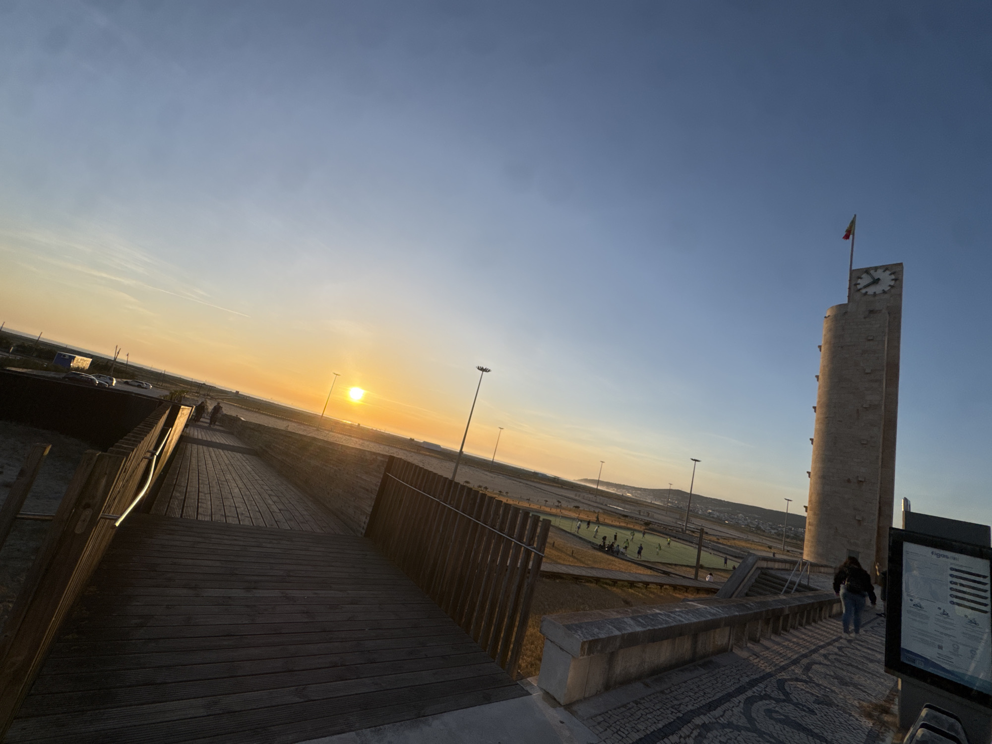 Captured in Figueira da Foz, Portugal, the image presents a boardwalk leading toward a beach at sunset. The sun casts a bright glow on the horizon, illuminating the surrounding area. A group of people are gathered on a nearby field, seemingly engaged in a soccer match. To the right stands a tall clock tower with a flag atop it, casting a shadow towards the walkway. A person is walking away from the tower along a paved walkway. An information board is positioned on the right of the image. The environment includes a combination of natural elements and man-made structures, capturing a blend of recreational and urban features.
