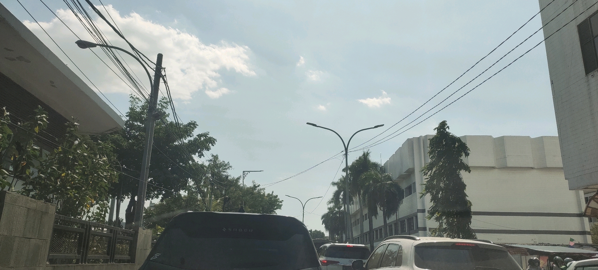 The image captures a street scene in Semarang, Indonesia, likely during daylight hours, as indicated by the bright sky with scattered clouds and the presence of streetlights that appear to be off. The perspective is from inside a vehicle, suggesting it is either parked or in traffic.
In the foreground, the rear of a dark-colored SUV is visible, with the word "SABER" printed on its tailgate. To the right, the front of a white SUV is partially visible. The street is lined with trees, including palm trees, and several cars are present, indicating traffic congestion.
Dominating the background is a large, modern-looking building with a distinct stepped or tiered facade, painted in a light color, possibly white or off-white. A building with a more traditional, lighter-colored facade and horizontal windows is visible on the far left. Electrical wires and multiple streetlights on poles crisscross the sky, a common sight in urban Indonesian environments.
The overall impression is a typical urban street environment in Semarang, characterized by a mix of modern architecture, natural elements like trees, and the presence of vehicles and infrastructure.
