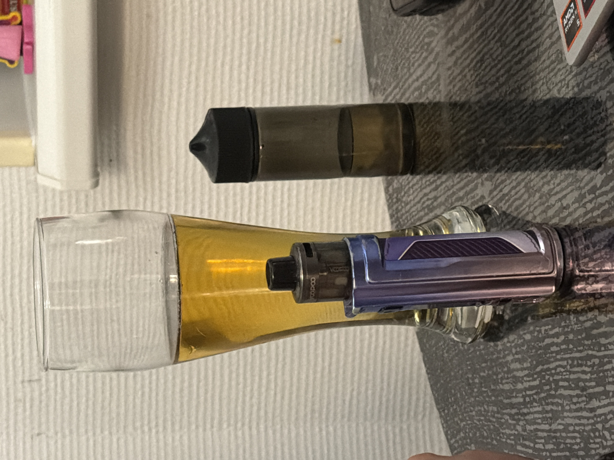 The image shows a still life arrangement featuring two vape devices and a glass with a golden liquid. In the foreground, a glass, possibly a shot glass or a small liqueur glass, contains a golden-amber colored liquid. Resting on the rim of this glass is a vape mod, a metallic device with blue and silver accents and a textured pattern. The vape mod is positioned horizontally across the glass, with its mouthpiece extending into the glass.
Behind the glass, a separate vape pod is visible. It is a dark, translucent cylindrical object with a black tip, lying horizontally. The background is a textured surface, appearing to be a tablecloth or similar fabric. To the left of the scene, a white cylindrical object and a small pink object are partially visible, suggesting other items on the surface. In the upper right corner, a corner of a laptop sticker with "AMD RYZEN 5" is visible.
The lighting in the image suggests an indoor setting, possibly with artificial light. There are no people visible, and the overall impression is that of a carefully arranged composition of vaping paraphernalia. The liquids and objects are clean and appear to be in good condition. The image does not convey any specific time of day or weather conditions, nor does it offer clear location cues beyond the presence of the AMD sticker. The emotions evoked are neutral, focusing on the objects themselves.