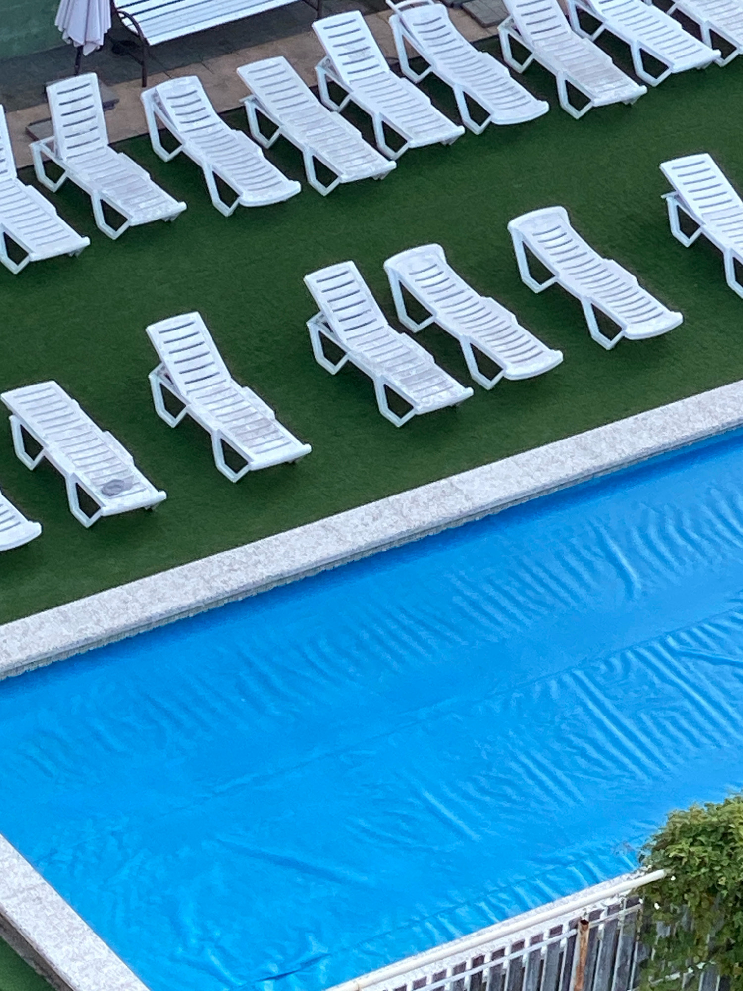 An overhead, high-angle shot shows a resort scene in Nebug, Russia, featuring rows of white plastic lounge chairs arranged on artificial turf adjacent to a swimming pool covered by a blue tarp. The chairs are empty and appear to be awaiting guests. A white railing and some greenery are visible in the lower right corner. The overall impression is one of quiet readiness.
