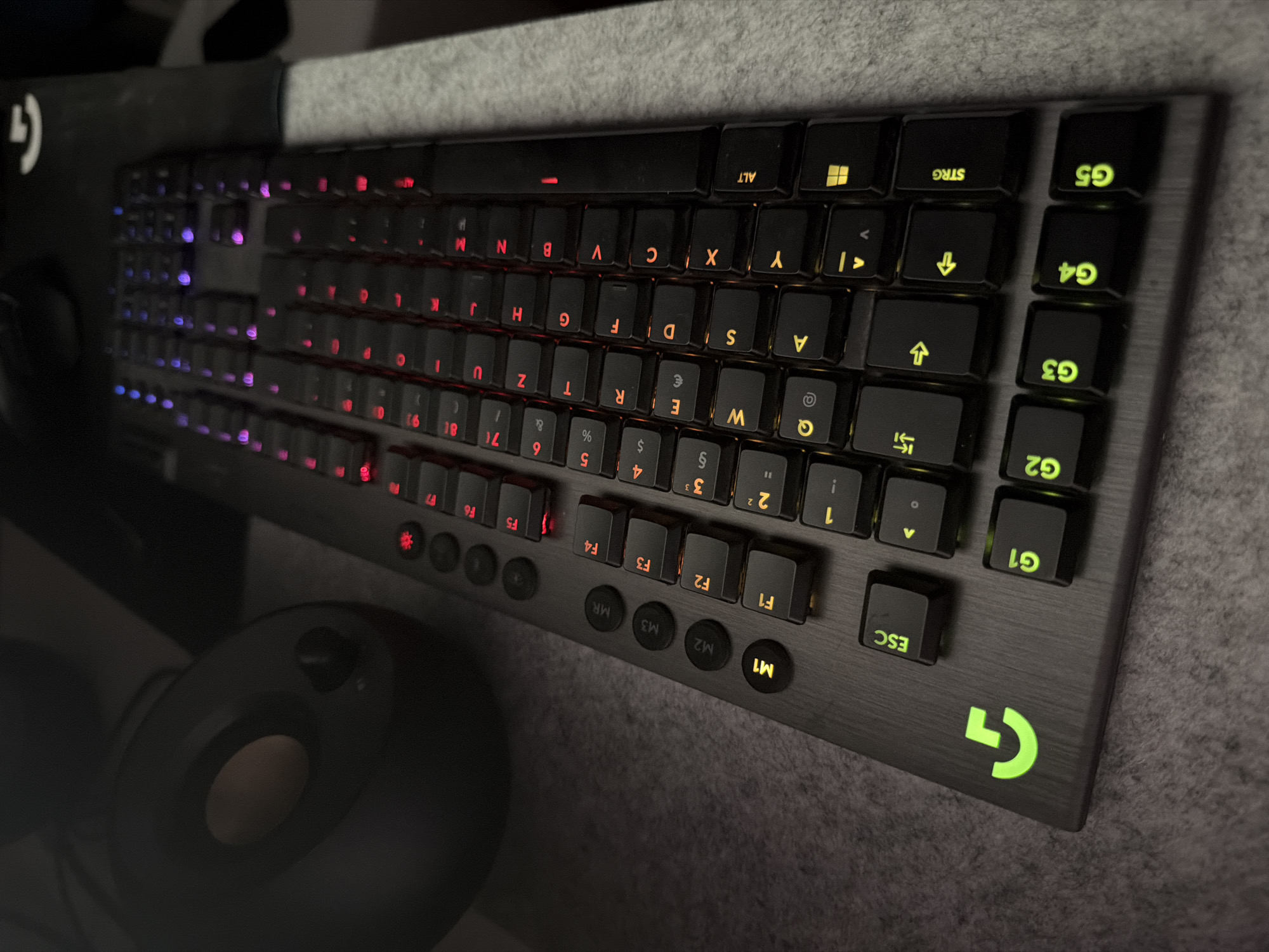 A Logitech G-series mechanical keyboard, with a brushed metal finish and black keycaps, is depicted in a close-up, top-down view. The keys are illuminated by a mixture of red and purple backlighting. Special function keys labeled G1 through G5 are visible on the left side, along with dedicated macro keys M1, M2, M3, and MR. The ESC key is also illuminated. To the left of the keyboard, a black speaker with a circular, tan-colored grill is partially visible. The keyboard and speaker rest on a textured gray surface, possibly a desk mat or cloth. The image is an indoor shot, likely taken in a gaming setup or office environment. There is no indication of the city of Esch-sur-Alzette, Luxembourg, or any people within the frame.