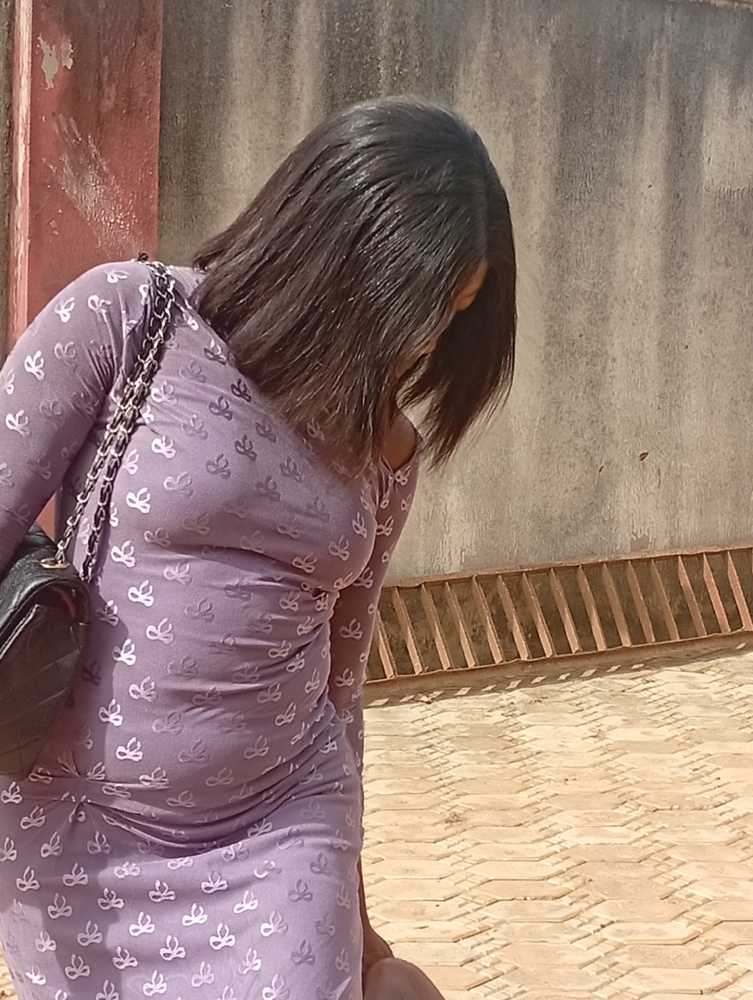 The photograph shows a woman in an unidentified city, but specifically in an urban environment identified as "Unknown, Unknown," wearing a light purple long-sleeved dress with a repetitive white pattern. The woman is positioned facing downwards, with her head tilted and dark hair obscuring her face. A black purse with a chain strap hangs off of her shoulder.
The background includes a wall with a red section on the left side and a gray, textured section to the right. Below the gray wall is a ledge with regularly spaced vertical slats. The ground is paved with light-colored hexagonal tiles.