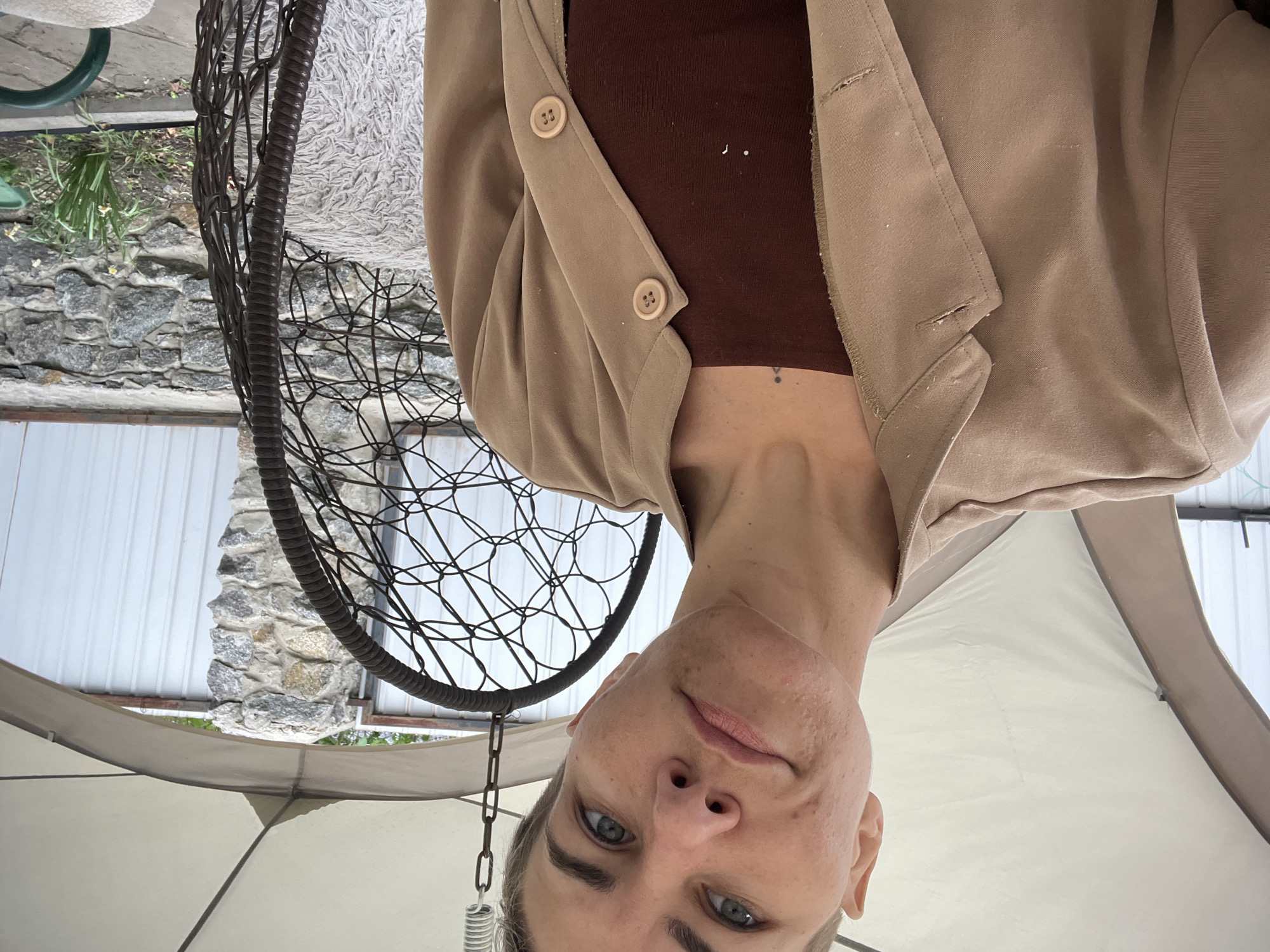 A woman with fair skin, blue eyes, and short gray hair is oriented upside down in the frame. She is wearing a brown top and a beige blazer with buttons. Behind her is a black, circular, hanging chair. In the background are elements suggesting an outdoor setting, including a white wall, a stone wall, and foliage. The setting appears to be under a tent or umbrella. There is no definitive evidence to confirm or deny the location is Boguchar, Russia.