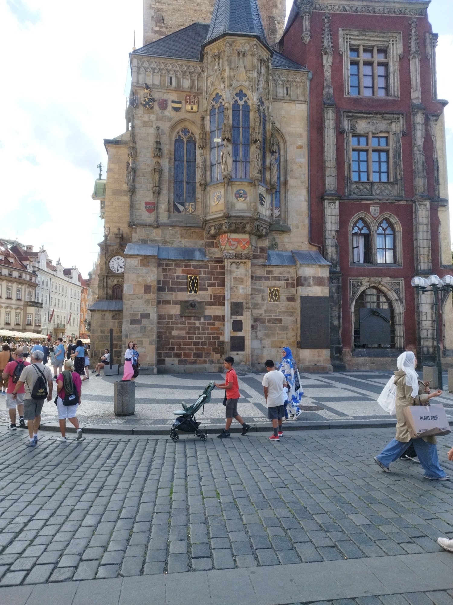 The image depicts a bustling outdoor scene in what appears to be a European city square. A large, ornate building with intricate stonework and Gothic-style windows dominates the background. Adjacent to it is a section of the building with a deep red facade, also featuring decorative windows. A large clock face is visible on the stonework of the older building.
Numerous people are walking on a cobblestone pavement in the foreground and mid-ground. They are dressed in casual summer clothing. Several individuals are carrying shopping bags. One person is pushing a stroller. The mood seems relaxed and leisurely, suggesting a typical day in a tourist area.
Notable details include the architectural style of the buildings, indicative of a historical European city. The time of day appears to be daytime, with bright lighting suggesting a sunny or partly cloudy sky. The ground is paved with textured cobblestones.
One visible object has text on it: a white shopping bag held by a person in the right mid-ground has the words "PLANT ARE" printed on it, although the full word is cut off. The overall scene conveys a sense of urban activity and historical charm.