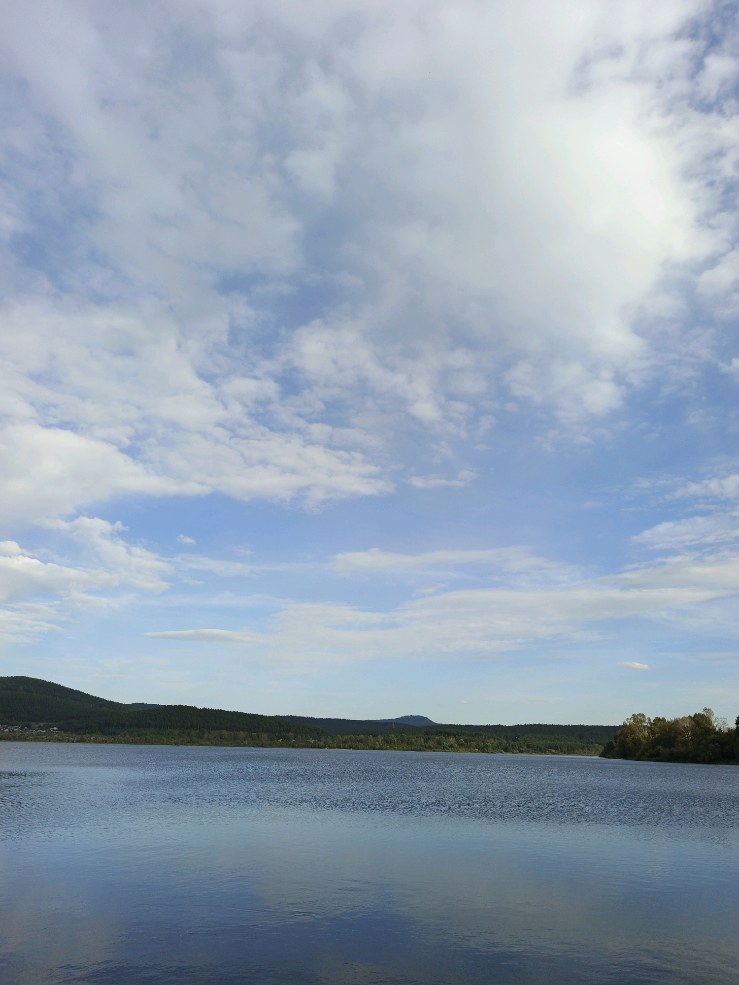 A wide, still body of water with a calm surface reflects the sky and distant treeline. The water occupies the bottom half of the frame. A dark green, rolling treeline with some visible structures of buildings is visible across the water, forming the horizon. Behind the treeline, a hill or mountain range is visible. The sky is a vibrant blue with numerous white, fluffy clouds scattered across it, some of which are dense and sunlit, while others are thinner and more dispersed. The overall scene is a natural landscape, likely a lake or large river, in a rural or forested area, and is possibly located in Zlatoust, Russia given the context provided. There are no people or animals visible in the image.