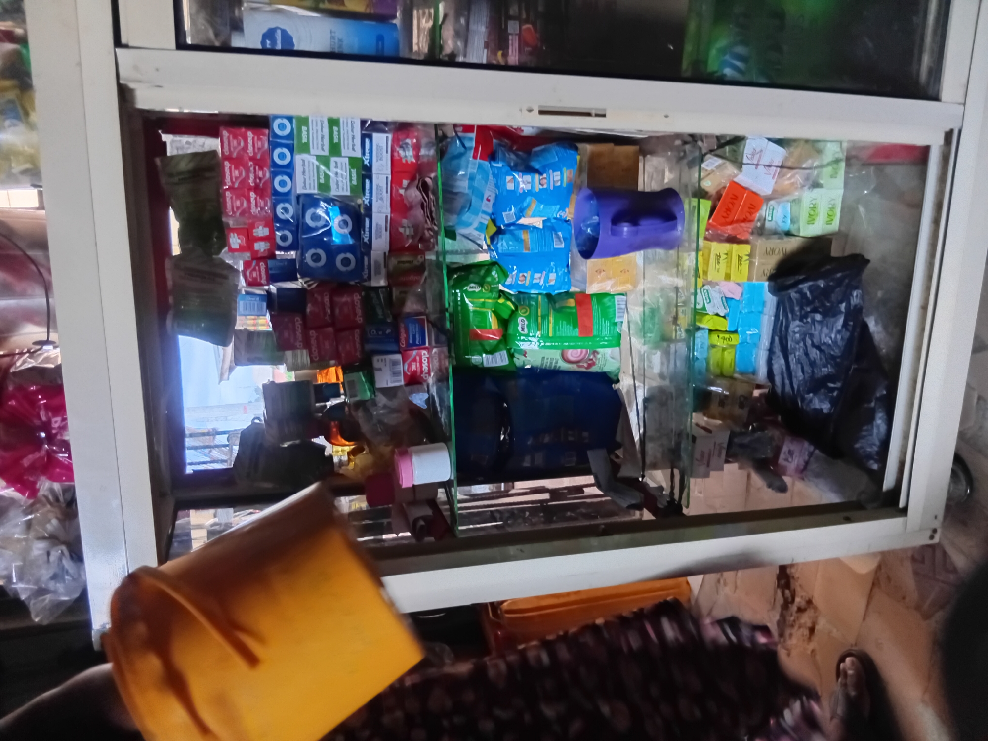 The image shows a display cabinet filled with various consumer goods, likely in a small shop. The cabinet has glass doors, allowing a clear view of the items inside. On the top shelf, there are packaged soaps and toothpaste. The middle shelf contains more packaged soaps, toothpastes, and possibly bags of snacks like "Milo." A purple mug is placed on this shelf, to the right. The bottom shelf displays more soaps, some in colorful packaging, and other small items. In the foreground, to the left, a person's arm and shoulder are visible, holding a bright yellow bucket. The background is blurred, suggesting the presence of other shelves or merchandise. The overall environment appears to be indoors, possibly a market stall or a convenience store.