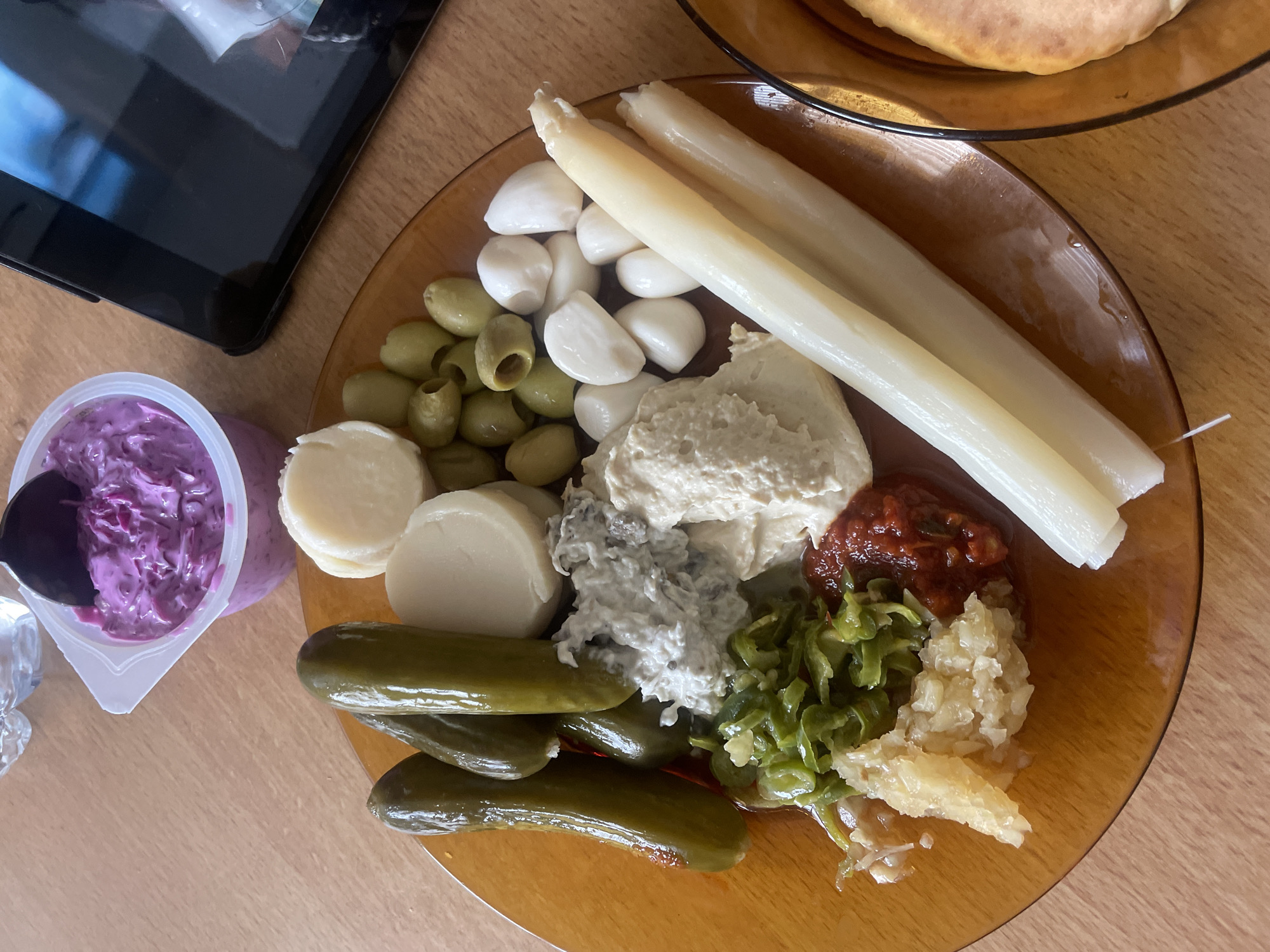 The image displays a meal set on a light-colored wooden table in Petah Tikva, Israel. The central object is an amber-colored glass plate containing various food items. On this plate, from left to right, are four whole dill pickles, several circular slices of a white cooked vegetable or soft cheese, a quantity of pitted green olives, and a cluster of small, white pickled garlic cloves or mozzarella pearls. Three long, straight, white spears, consistent with white asparagus or pickled hearts of palm, are positioned along the right edge of the plate. Five distinct condiments are centrally arranged: a creamy greyish-white dip (possibly fish-based or labneh), a beige hummus, a mound of finely chopped green pickled vegetables, a dollop of chunky reddish sauce (e.g., matbucha or harissa), and a pile of finely chopped yellow preserved lemon. To the upper right of the main plate, a small clear plastic container holds a vibrant purple red cabbage salad, with a black spoon partially submerged. A black tablet, enclosed in a dark case, is partially visible on the right, displaying a light-colored image on its screen. A second, partially visible amber-colored glass plate on the far right holds a piece of flatbread, likely pita. A portion of a clear glass or plastic cup is visible in the upper left background. No people are present in the scene.