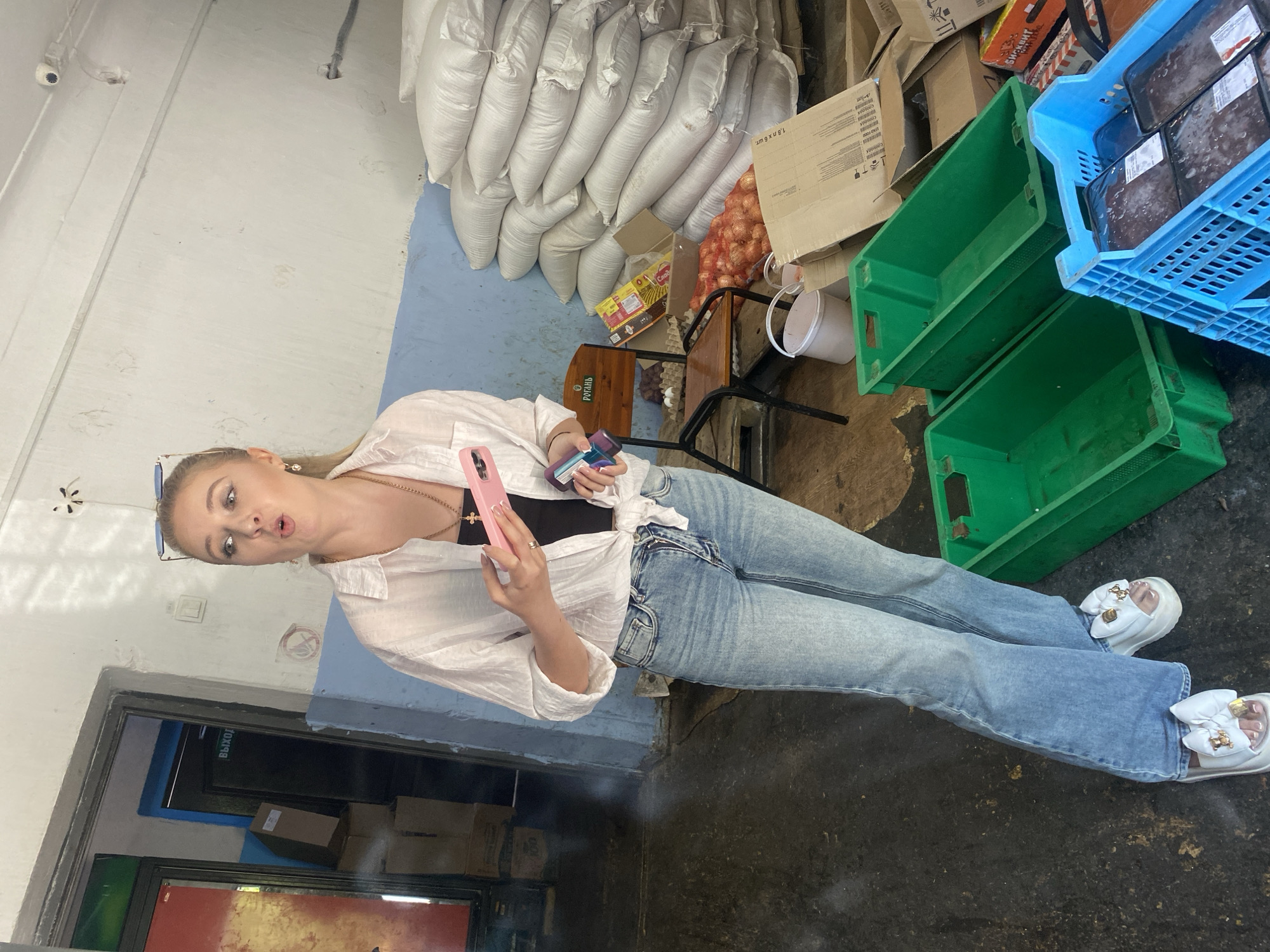 A young Caucasian woman with blonde hair pulled back and wearing sunglasses on her head stands in a dimly lit warehouse-like room. She is wearing a white open-fronted shirt tied at the waist over a black top, and light blue flared jeans. She is holding a pink smartphone and another object in her hands. The floor is concrete and appears dusty. To her right, there are stacks of large white sacks, possibly containing grain, and piles of onions. In the foreground, there are green and blue plastic crates, with one blue crate containing packaged items. A wooden chair is visible behind her, and a doorway with Cyrillic text is in the background. The room has blue and white painted walls.