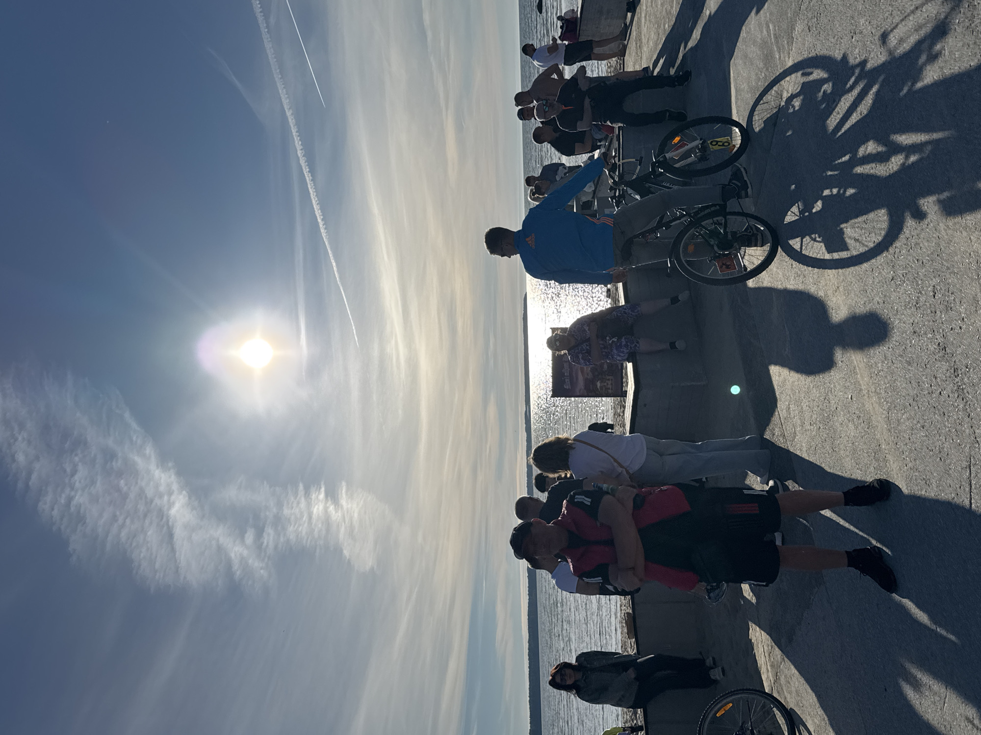 This image captures a group of people gathered by the sea on a sunny day. The sun is high in the sky, creating a bright glare on the water. The scene appears to be a public promenade or pier, with a paved walkway in the foreground and a body of water in the background. Several individuals are standing and sitting, some are looking towards the sea, while others are interacting with each other. Two bicycles are visible, suggesting the possibility of a cycling event or recreational activity. The sky is partly cloudy with some wispy clouds and contrails from an airplane. The overall atmosphere is relaxed and outdoor-oriented.
The location is the Tallinn Sea Promenade in Tallinn, Estonia. The distinctive architecture of the buildings in the distance, the style of the promenade, and the visible signage (though not legible in detail) are characteristic of this area in Tallinn. The presence of the sea and the specific type of sky also aligns with the geographical and meteorological conditions typical of the Baltic Sea coast in Estonia.