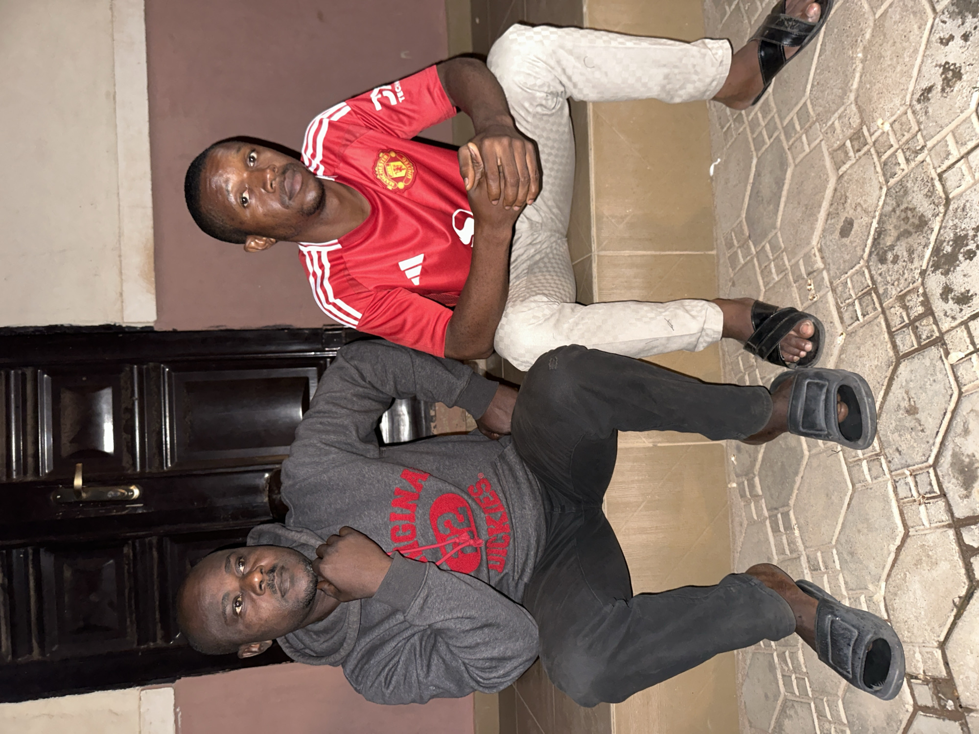 Two adult males are seated on a concrete step in front of a dark wooden door with a brass handle. The male on the left wears a grey hooded sweatshirt with red lettering that says "ORIGINAL 2dickIES" and dark grey jeans. He has his chin resting on his hand. The male on the right wears a red Manchester United jersey with white stripes on the sleeves and light-colored checkered pants. His hands are clasped in his lap. Both men are wearing dark-colored slides. The ground in front of the step is tiled with a patterned design. The overall lighting suggests it is nighttime. This scene is located in Narayi, Nigeria.
