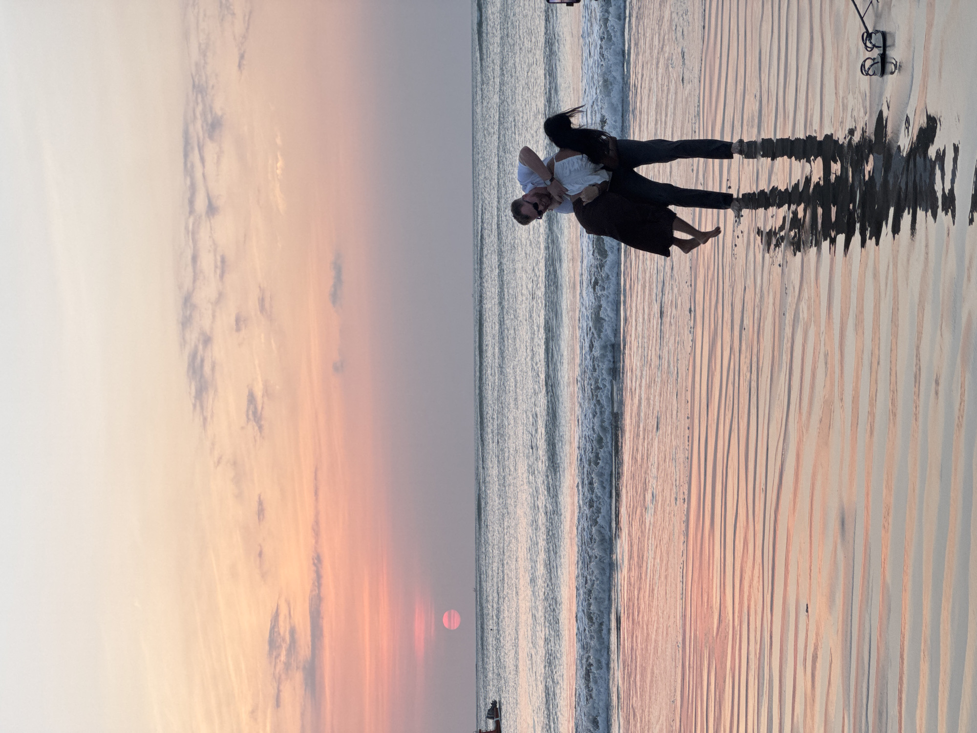 The image depicts a romantic moment on a beach in Kuta Selatan, Indonesia. A man is seen lifting a woman in his arms, as if dancing, on the wet sand near the ocean's edge. The sunset casts a warm glow over the scene, with the sky displaying hues of pink, orange, and pale yellow. Gentle waves are rolling onto the shore, creating ripples in the sand and reflecting the colors of the sky. In the background, the ocean stretches towards the horizon, with the sun, a distinct pink circle, visible above it. A small boat is present on the left side of the image, near the horizon line. A pair of sandals rests on the sand in the foreground, to the right of the couple. The overall atmosphere is serene and picturesque, capturing a moment of intimacy against the backdrop of a beautiful sunset.