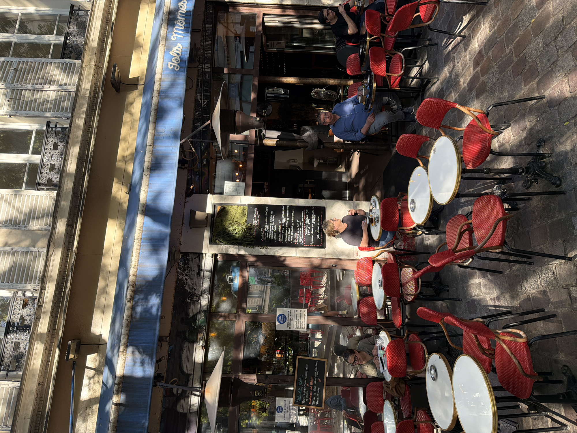 The image depicts the exterior of a Parisian cafe called "Jolis Mômes." Several people are seated at red chairs and small round tables on a stone-paved street, enjoying drinks or meals. The cafe has a blue awning and a menu board displayed outside. Reflections in the glass windows show the interior and street scene. The architecture suggests a traditional Parisian building. Shadows indicate it is daytime. The cafe is located in Paris, France.