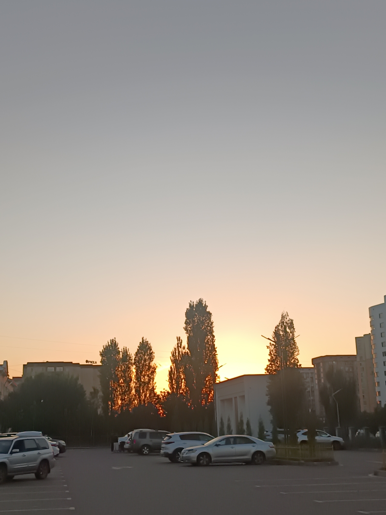 The image shows a parking lot in Bishkek, Kyrgyzstan, during sunset. Tall trees are silhouetted against the orange glow of the setting sun, with several buildings visible in the background. Cars are parked in the lot, some of which are silver SUVs and sedans. The sky is a gradient of pale gray to soft orange. The scene appears to be an outdoor urban environment.