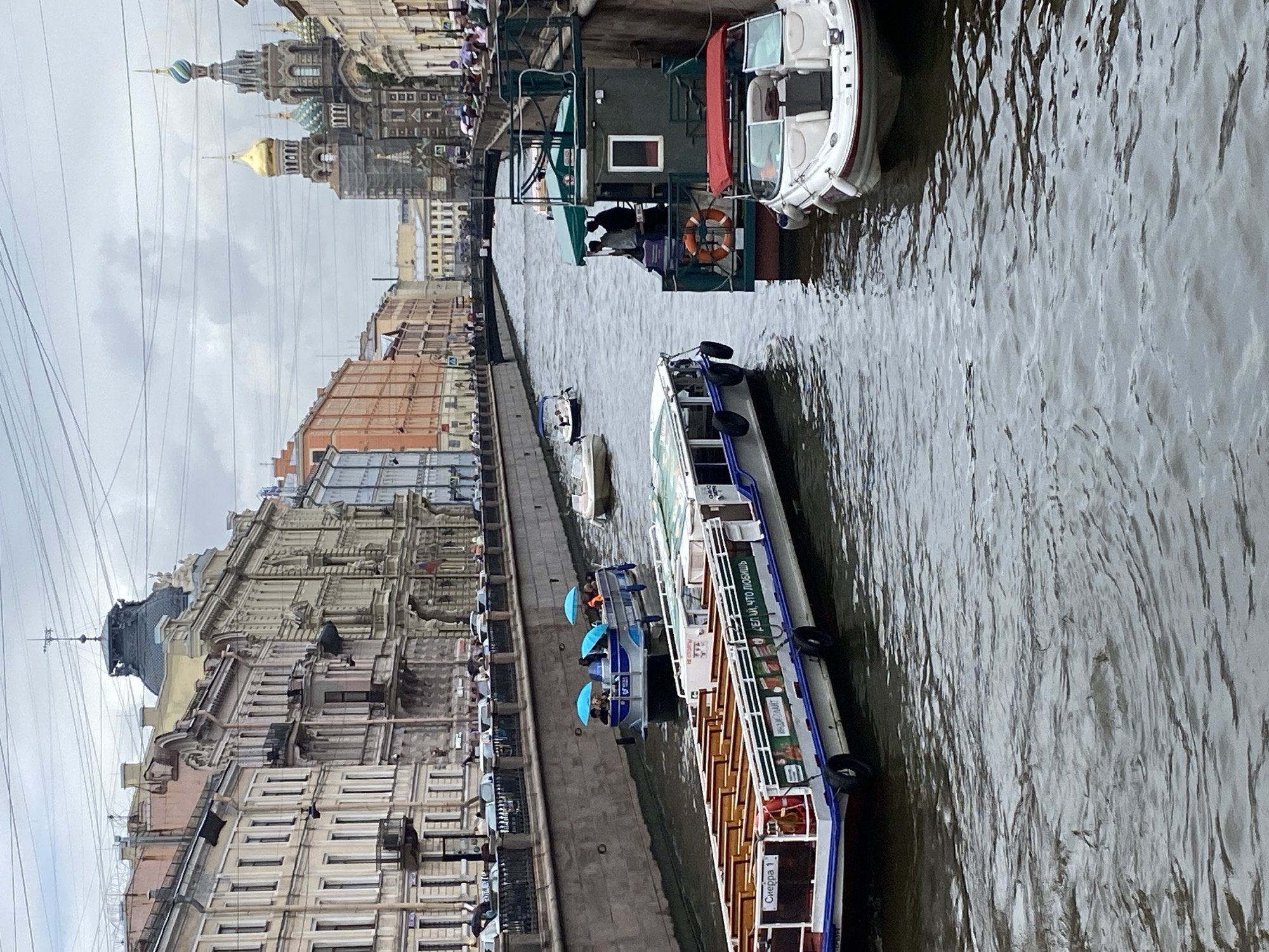 The image captures a canal in Saint Petersburg, Russia, likely the Griboyedov Canal, given the distinctive architecture and the presence of the Church of the Savior on Spilled Blood in the background. The canal is flanked by multi-story, ornate buildings with classical facades. Several boats are navigating the waterway. In the foreground, a long tour boat with many empty seats is docked or moving slowly, its side displaying advertisements in Cyrillic. To its right, a smaller, darker boat with a green canopy serves as a smaller vessel, possibly for a tour guide or maintenance, with at least two people standing on its deck. Further up the canal, a blue and white tour boat carries passengers who are holding up blue umbrellas. In the distance, the iconic onion domes and spires of the Church of the Savior on Spilled Blood are visible, under a cloudy sky. The water surface is rippled, indicating a moderate flow or presence of multiple boats. Along the canal walls, people are visible walking or standing, some holding umbrellas, suggesting a possibly rainy or overcast day.
