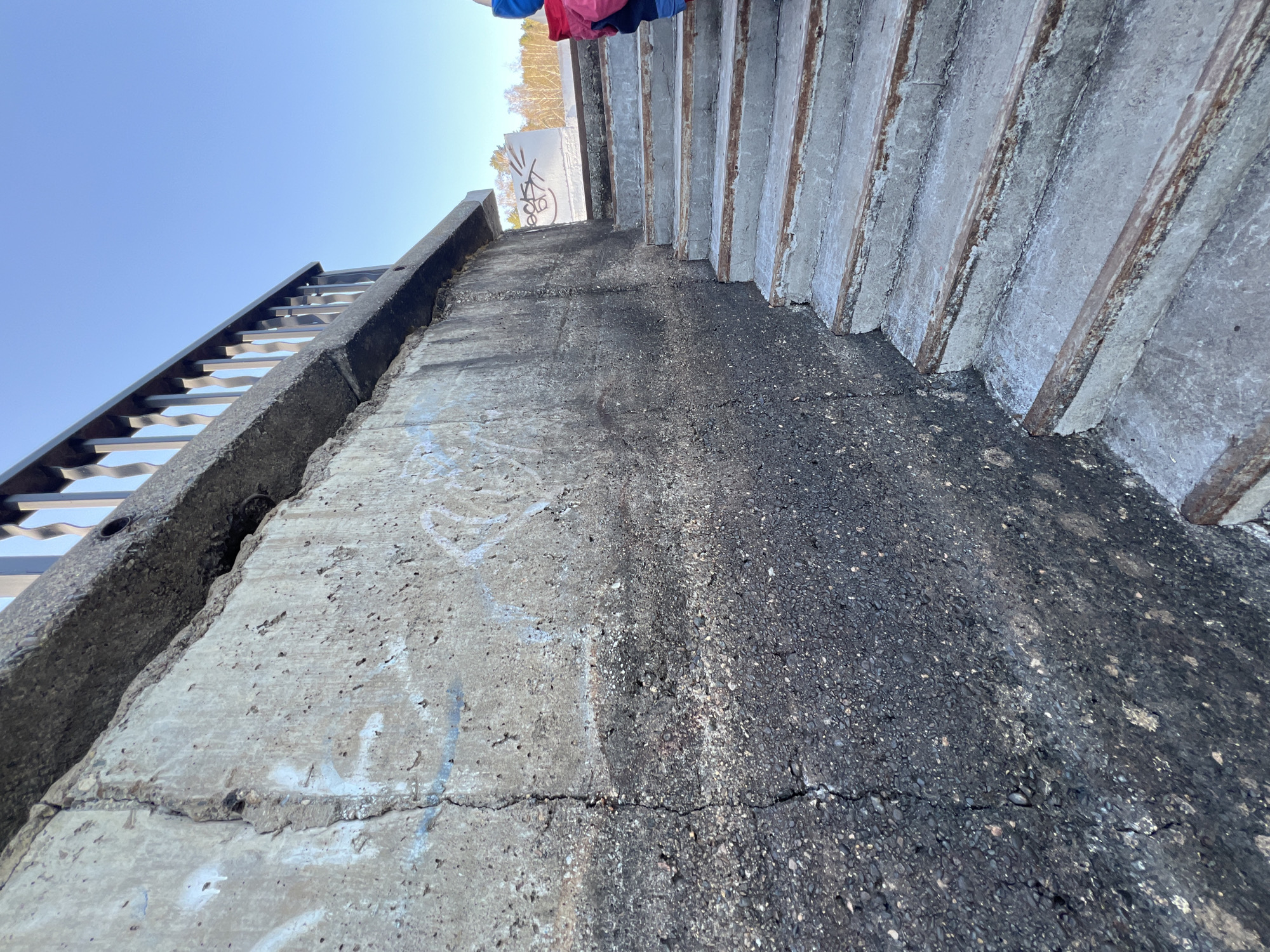 The image shows a low-angle view of a concrete staircase and adjacent walkway in Chaikovsky, Russia. The staircase is to the right of the image frame with the bottom of the staircase being the lowest part of the image frame. The walkway and railing are to the left. Graffiti is visible on the staircase wall. There is cracking along the base of the concrete. The sky is visible in the upper left corner of the frame. At the top of the image frame, garments appear, hanging from the top of the staircase.
