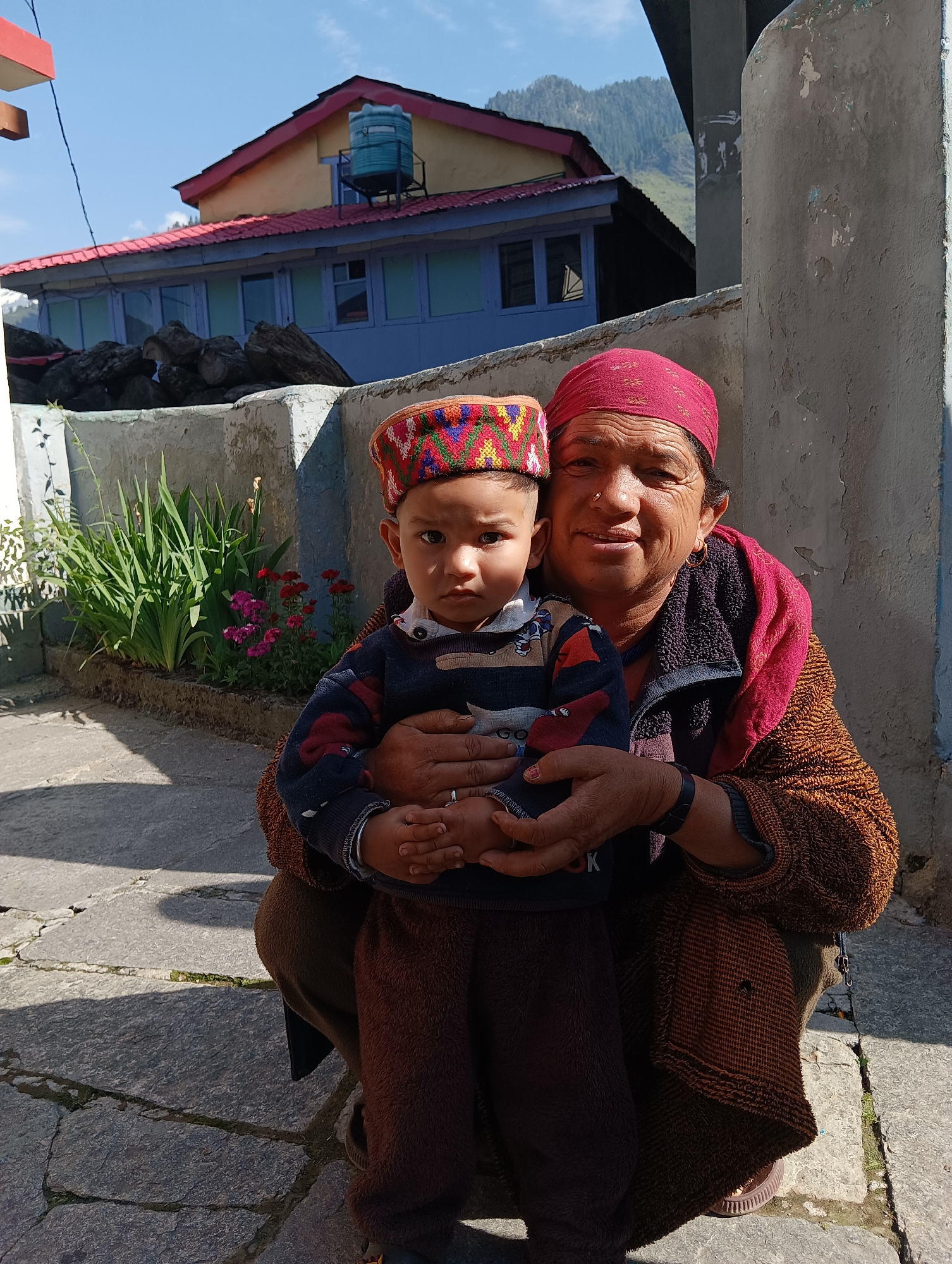 In the city of Kullu, India, an elderly woman with a red headscarf and a brown knitted shawl is holding a young child. The child is wearing a colorful, traditional cap and a dark sweater with a striped pattern. They are positioned in front of a stone wall, with a planter box filled with greenery and pink flowers to the left. In the background, a building with a red roof and light blue walls is visible, along with a water tank on its roof. The scene is bathed in sunlight, casting shadows on the stone-paved ground. The woman is smiling, and her gaze is directed towards the viewer, while the child's gaze is also towards the viewer. The woman's right arm is around the child, and her left hand is gently holding the child's hands.