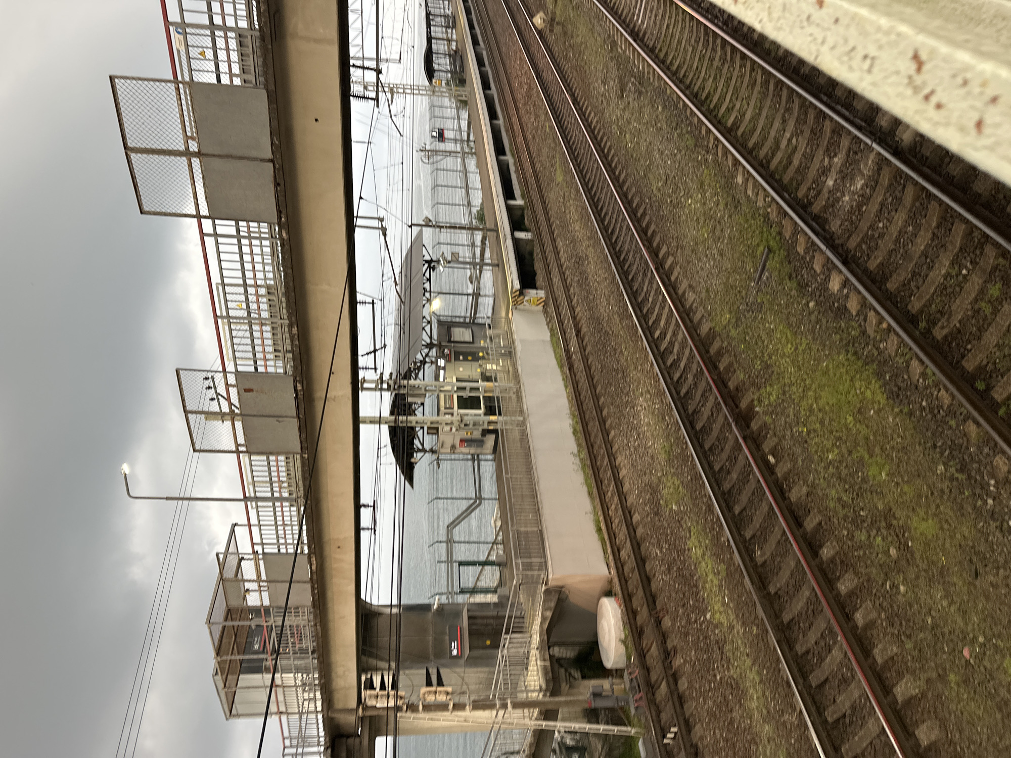 The image depicts a train station located in Sochi, Russia, specifically the Adler train station. The scene is captured on an overcast day, with a dramatic sky filled with gray clouds. In the foreground, multiple sets of train tracks stretch into the distance, bordered by gravel and patches of green grass. A concrete walkway or platform is visible on the right side of the frame, partially obscuring the tracks.
Above the tracks, a modern overpass is visible, featuring a pedestrian walkway with metal railings and fencing. A street lamp is mounted on the overpass, casting a glow into the scene. On the left side of the overpass, a small enclosed structure with a metal frame and mesh walls can be seen. Further along the overpass, additional fencing and platforms are present.
Beyond the train station infrastructure, the Black Sea is visible, its waters appearing gray and choppy under the cloudy sky. A fence runs along the edge of the platform overlooking the sea. The overall impression is one of a functional, somewhat stark, but functional transportation hub with a backdrop of natural scenery. There are no people visible in the image.