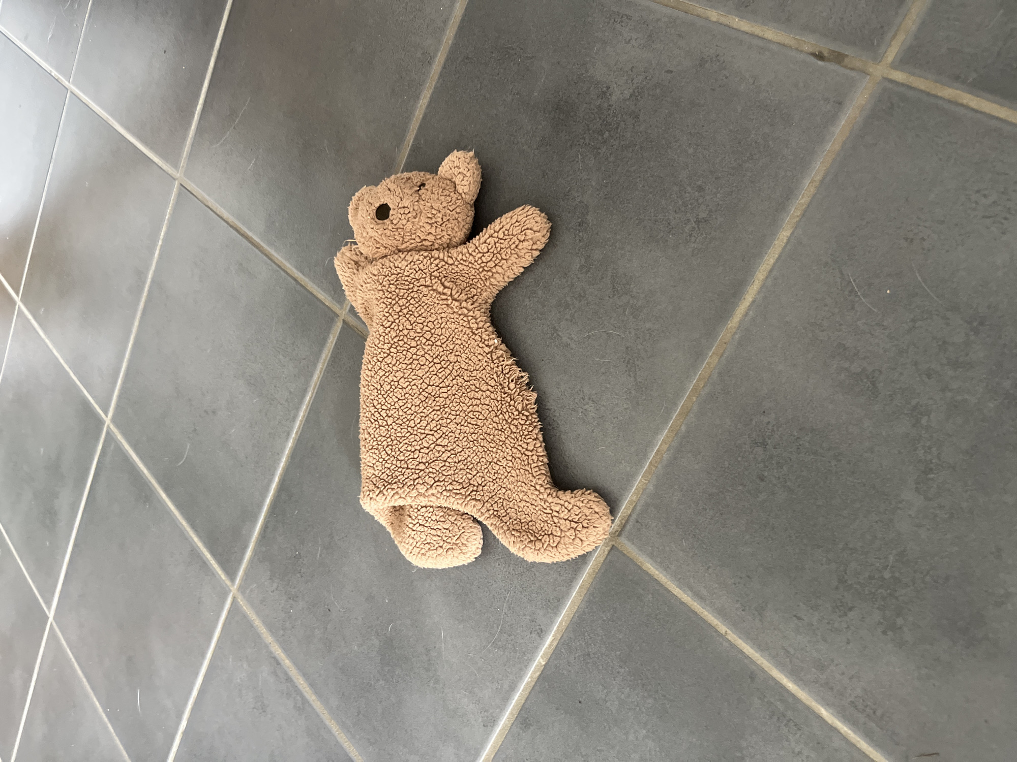 A brown, fuzzy stuffed bear lies on a grey tiled floor. The bear is lying on its stomach with its legs and arms spread out. Its head is turned to the right. The tiles are square and laid in a grid pattern with grout lines. There are no people visible in the image. The lighting suggests an indoor environment. The scene is devoid of any context that would indicate its location within Gertwiller, France.