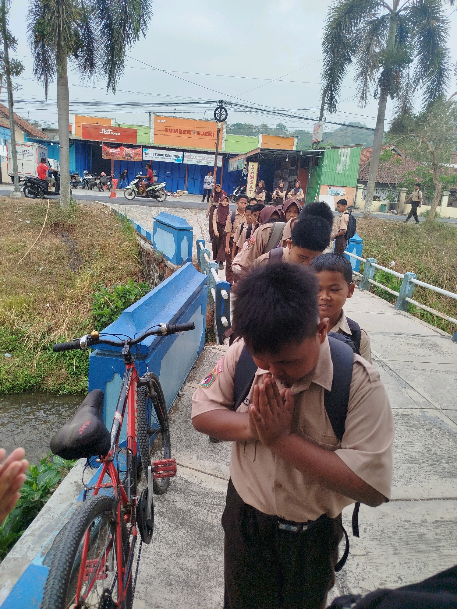 In Kedungwaru, Indonesia, a group of male students in tan uniforms and dark trousers, some wearing backpacks, are walking on a concrete bridge over a small canal. The student in the foreground is bowing with hands clasped in front of him. A red bicycle with a black seat is leaning against the blue railing of the bridge. In the background, there are buildings with signs including "J&T EXPRESS", "SUMBER REJEKI", and "SAMSAT BUDIMAN". Several motorbikes are parked in front of the buildings. Palm trees are visible on both sides of the scene. The sky is overcast.