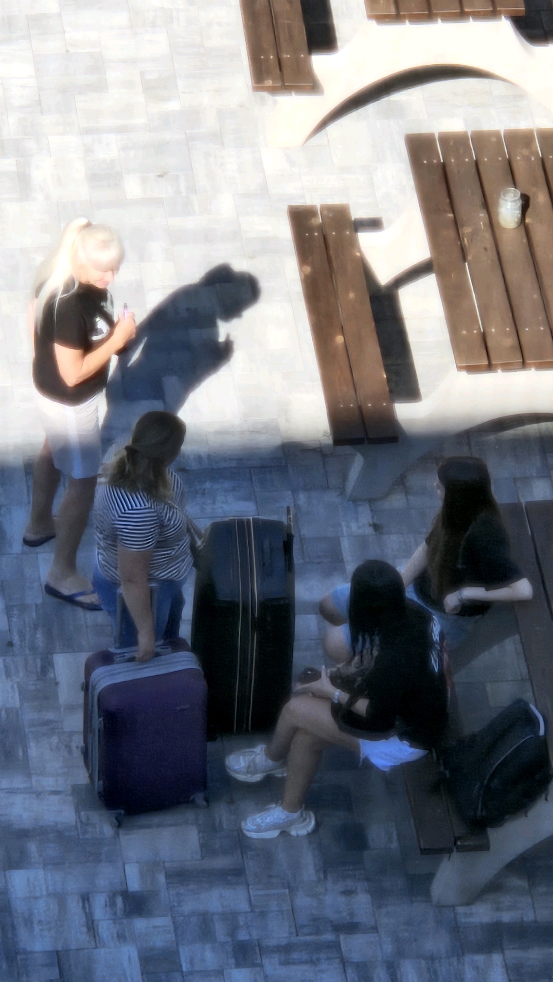 An overhead shot captures a group of four individuals standing and sitting on a stone patio next to wooden picnic tables. The scene appears to be outdoors, possibly at a park or recreational area.
On the left, a woman with blonde hair in a ponytail wears a black t-shirt and grey shorts. She is holding a pink object, possibly an e-cigarette or vape. Next to her, a shadow suggests another person, but they are not clearly visible.
In the middle ground, a woman with brown hair tied back in a ponytail wears a striped t-shirt and jeans. She is standing next to a large black suitcase and appears to be interacting with a purple suitcase in front of her.
In the foreground and right side, two individuals are seated. One, with long dark hair, wears a black t-shirt and white shorts, with white sneakers. They are seated on a wooden bench, and a black backpack is placed behind them. The other individual, partially visible, wears a blue t-shirt and also has dark hair.
The stone patio is paved with rectangular tiles in varying shades of grey. The wooden picnic tables are weathered and appear to be located in a sunny area, casting distinct shadows. A small glass jar is visible on one of the tables. The overall lighting is bright, suggesting daytime. The location is specified as Prešov, Slovakia.