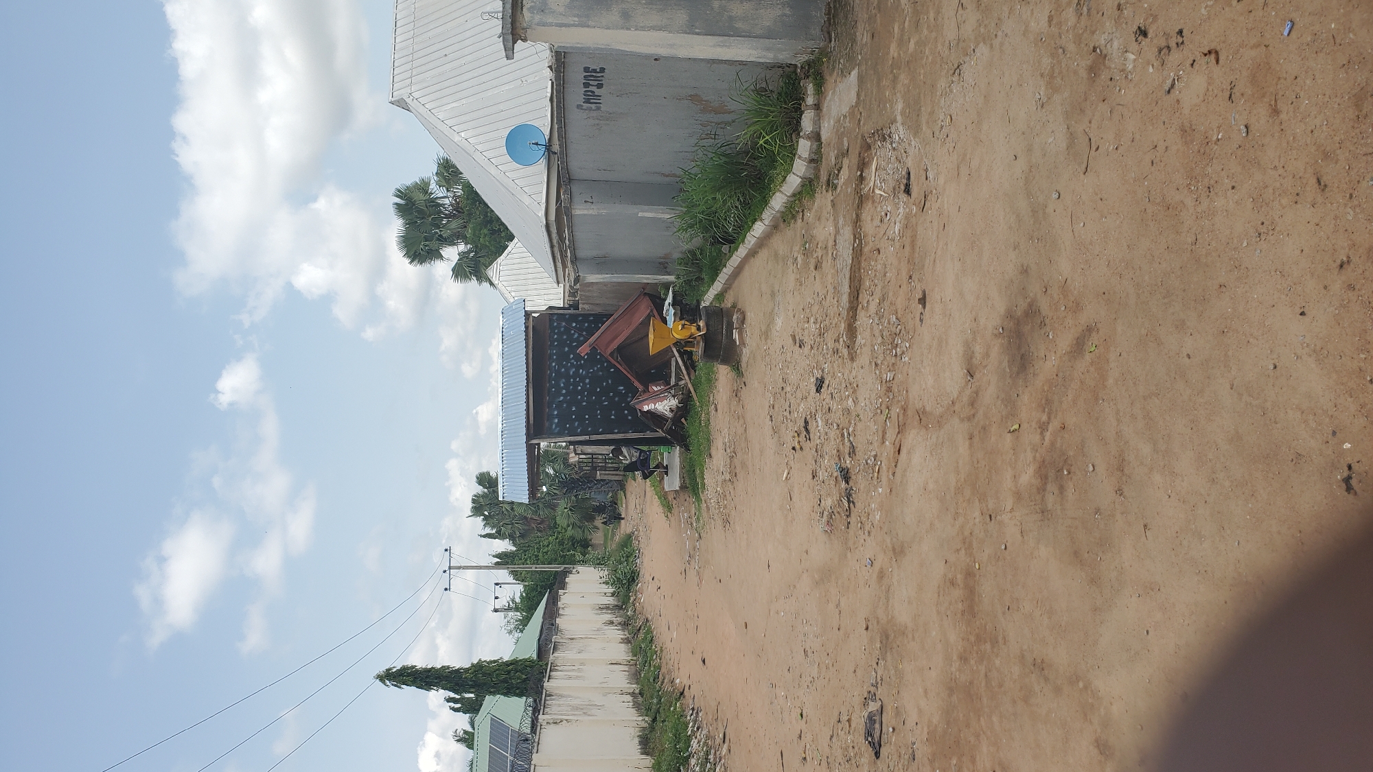 The image depicts an unpaved road in a town, with residential buildings and fences lining its sides. On the left, a beige concrete wall runs along the property line, with solar panels visible on the roof of a building behind it. A tall, slender tree stands near the wall. On the right, a gray concrete wall is visible, with the word "EMPIRE" spray-painted in black. A blue satellite dish is mounted on the roof of a building behind this wall.
In the middle of the road, there is a small, open-sided structure with a corrugated metal roof. Against this structure, a person is seated on a low ledge, facing away from the camera. Several objects are piled up around this structure, including what appears to be a red metal frame, possibly from a piece of machinery, and a yellow device mounted on tires, resembling a small mill or grinder. The road itself is made of compacted dirt and gravel, with patches of dry grass and scattered debris. In the distance, a few people are walking along the road. The sky is partly cloudy with a mix of white and gray clouds against a pale blue backdrop. The overall scene suggests a somewhat undeveloped or rustic urban environment.