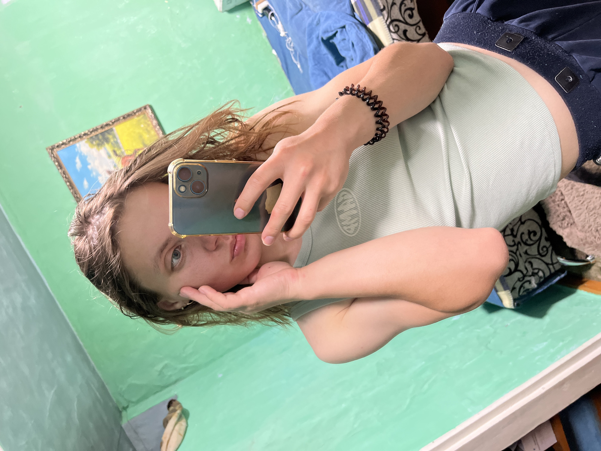 The image shows a young person taking a mirror selfie in what appears to be a bedroom. The person is holding a smartphone in their right hand, angled to capture their reflection. They are wearing a light green ribbed tank top and dark blue bottoms. Their left hand is touching their face, near their cheek and temple. A dark, coiled bracelet is visible on their right wrist.
The background is a brightly painted green wall. To the left, a framed picture of a landscape with a blue sky and yellow field hangs on the wall. To the right, a bed with blue bedding is partially visible. There is a shelf or furniture visible on the far left with some items on it. The lighting suggests indoor illumination.
There is no information within the image to suggest that this location is in Nizhny Tagil, Russia.