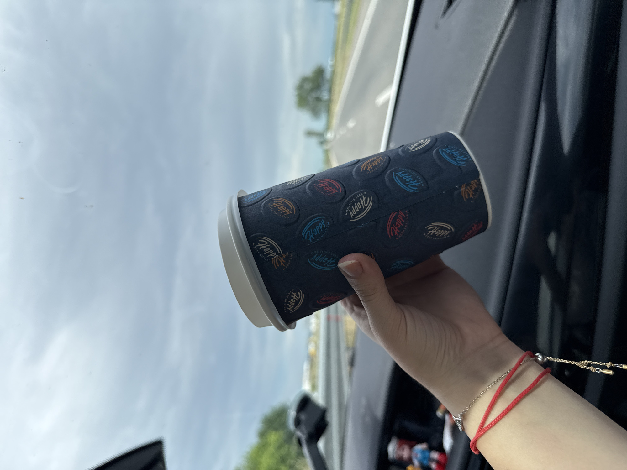 A person's hand holds a dark-colored disposable coffee cup inside a car, likely in Radinovo, Bulgaria. The cup is adorned with a repeating pattern of circular logos in various colors (blue, red, orange, beige) featuring the word "Happy" and the phrase "KEEP SMILING." The cup has a white rim. The hand, wearing two bracelets—a red string bracelet and a silver-toned chain bracelet with a small charm—is positioned in the lower right of the frame.
The setting appears to be the interior of a vehicle, with parts of the dashboard and windshield visible. Through the windshield, a bright, hazy sky is visible, indicating daytime. A blurred outdoor scene suggests a road or highway with distant trees and potentially a field. The overall impression is that of a journey or a stop along a route. The image is taken at eye level, looking out from the car.