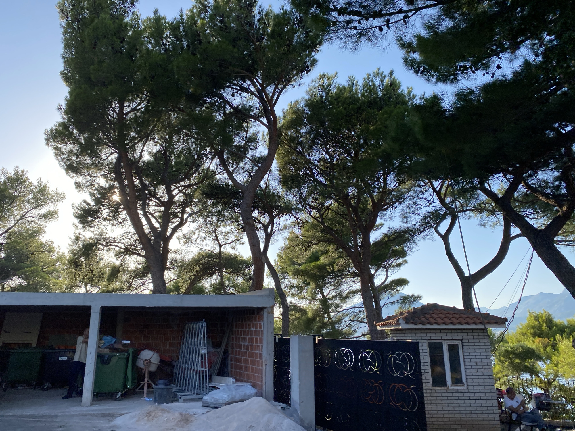 This image captures a scene in the city of Bar, Montenegro, featuring an outdoor area under a clear blue sky. Dominating the upper portion of the frame are several large pine trees with their dense green foliage and visible pinecones. Sunlight filters through the branches, creating dappled lighting.
In the foreground, there is a covered structure with a concrete roof, open on one side, revealing brick walls. Inside, several large green dumpsters are visible, along with a concrete mixer and scaffolding. A person, wearing a light-colored shirt and dark pants, stands near one of the dumpsters.
To the right of the covered structure is a small, white brick building with a tiled roof and two windows. Adjacent to this building is a black, ornate metal gate. Further to the right, two individuals are seated outdoors, with one person in a white shirt and another partially visible, near a table and chairs. The background shows a body of water and distant mountains, indicating a scenic coastal or lakeside location. The ground in front of the structures appears to be a mixture of sand and gravel, suggesting ongoing construction or landscaping work.