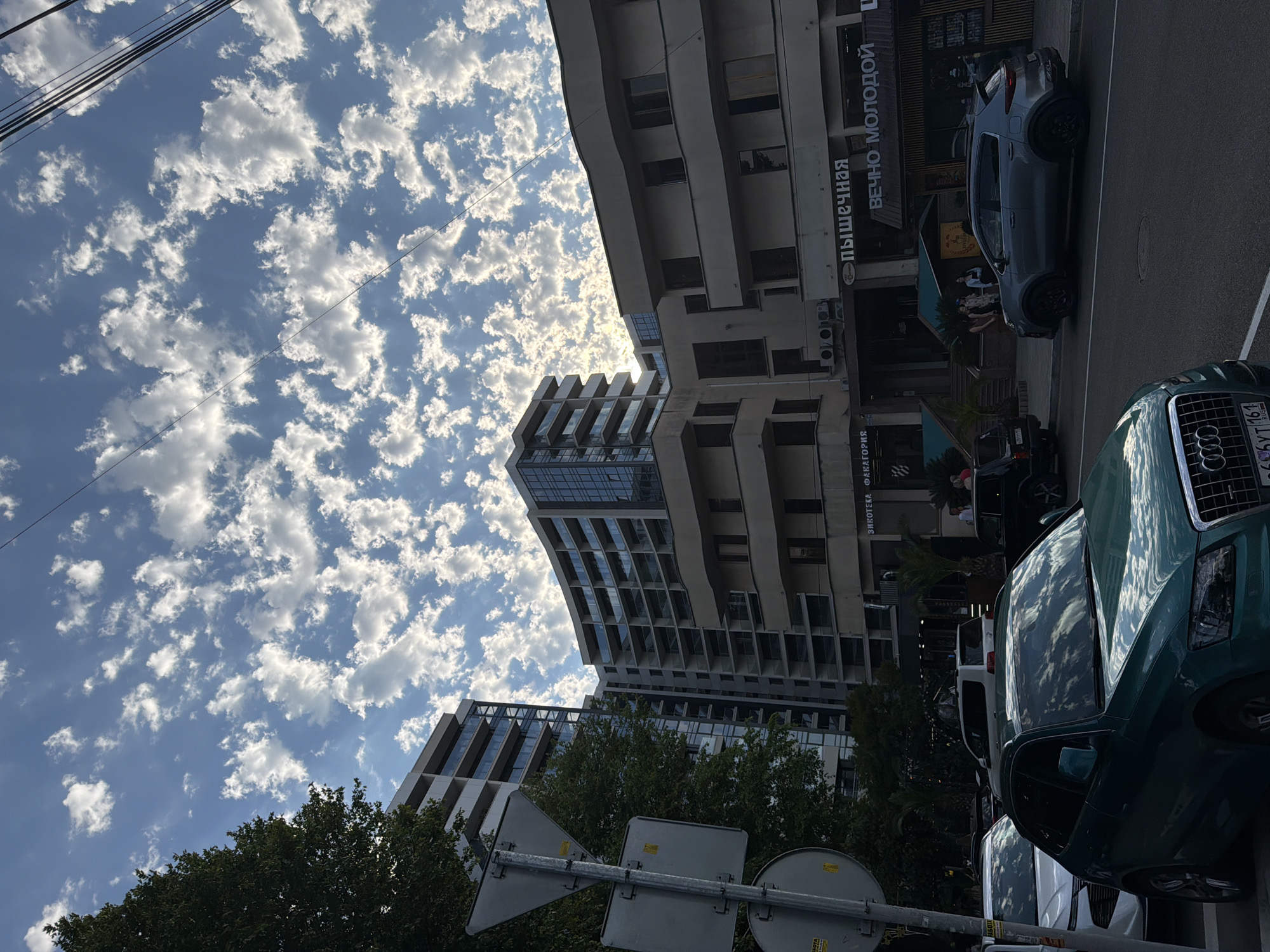 This media file depicts a street scene in Sochi, Russia, characterized by a bright, cloudy sky and modern architecture. The image is oriented vertically, with the sky occupying the upper left portion and the street and buildings on the right.
Dominating the sky are numerous cumulus clouds scattered across a vibrant blue expanse, suggesting a sunny day with scattered cloud cover. Several utility wires are visible stretching across the sky.
In the foreground on the right, a dark green Audi SUV is parked on the street. Its glossy surface reflects the sky and surrounding environment. Further down the street, other vehicles, including a white van and at least one other car, are also visible.
The background features multi-story buildings with contemporary designs, featuring numerous windows and balconies. Some buildings appear to be residential or mixed-use, with commercial storefronts at street level. One building has signage with Cyrillic text. Visible text includes "ПИЩЕВЫЯ" and "ВЕЧНО МОЛОДОЙ".
The scene suggests a typical urban environment, possibly during daytime, given the bright lighting and the appearance of the sky. The presence of parked cars and buildings indicates a populated area. No specific activity or event is clearly depicted, other than the static presence of vehicles and structures. The overall mood is calm and ordinary.